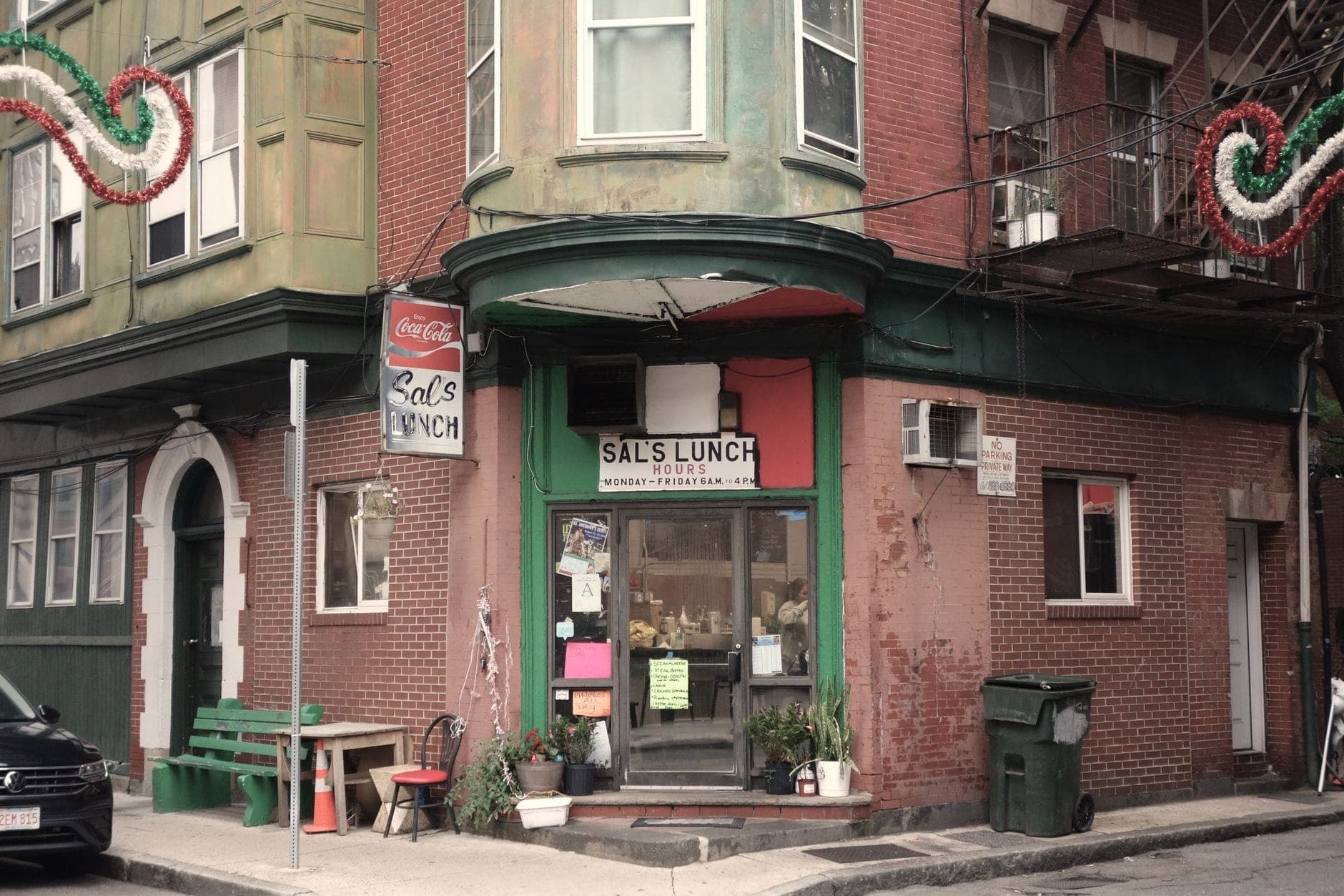 Vue de la rue de Sal's Lunch, un modeste restaurant de coin dans le North End de Boston, avec des enseignes, des bancs et des plantes à l'extérieur.