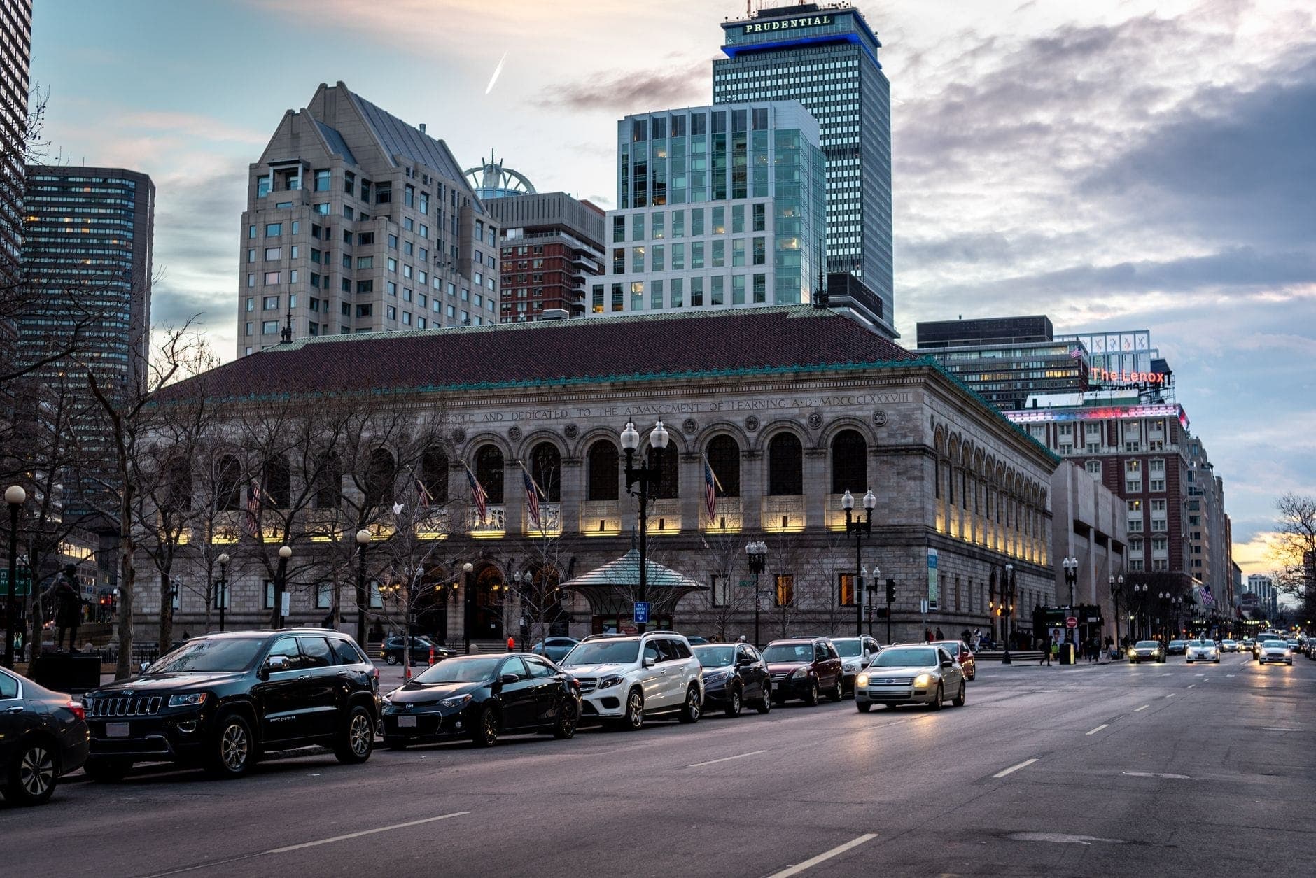 Vue de la majestueuse façade Renaissance Revival de la Boston Public Library au crépuscule, encadrée par les gratte-ciel modernes de la ville sous un ciel de feu.