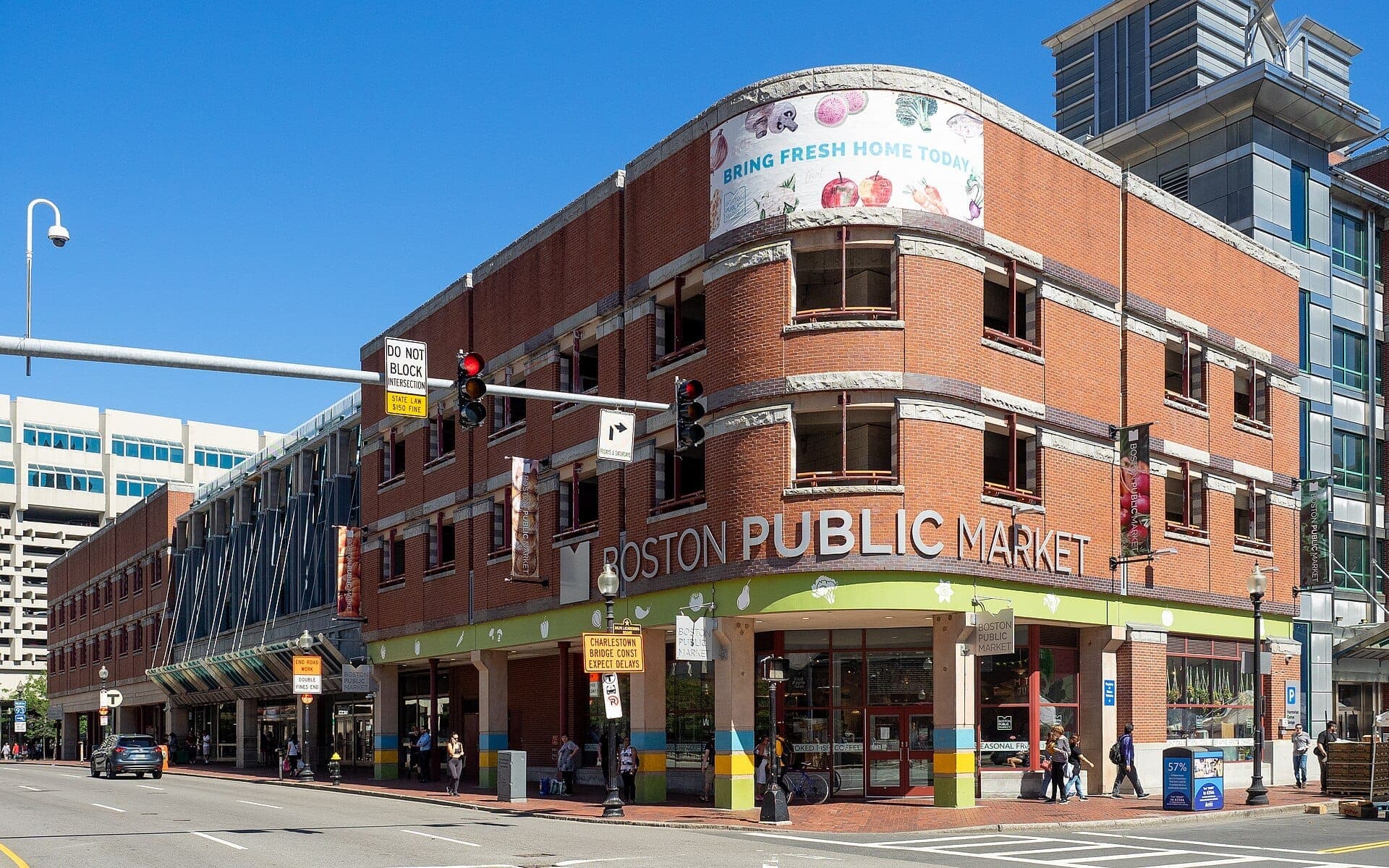 Bâtiment en brique du Boston Public Market avec de grandes fenêtres et une signalétique colorée par une journée ensoleillée, situé dans une rue animée de la ville.