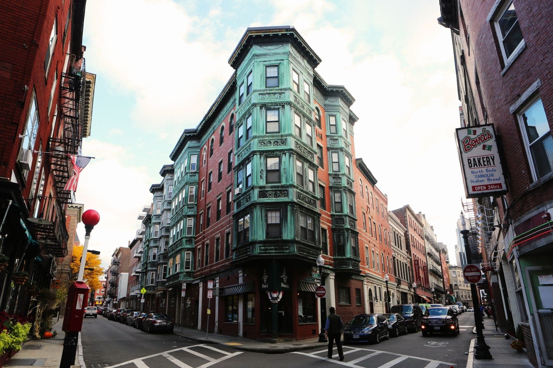 Scène de rue à Boston dans le North End, montrant des bâtiments classiques en brique, l'enseigne d'une boulangerie au coin de la rue et des passants, soulignant la facilité de déplacement à pied dans la ville.