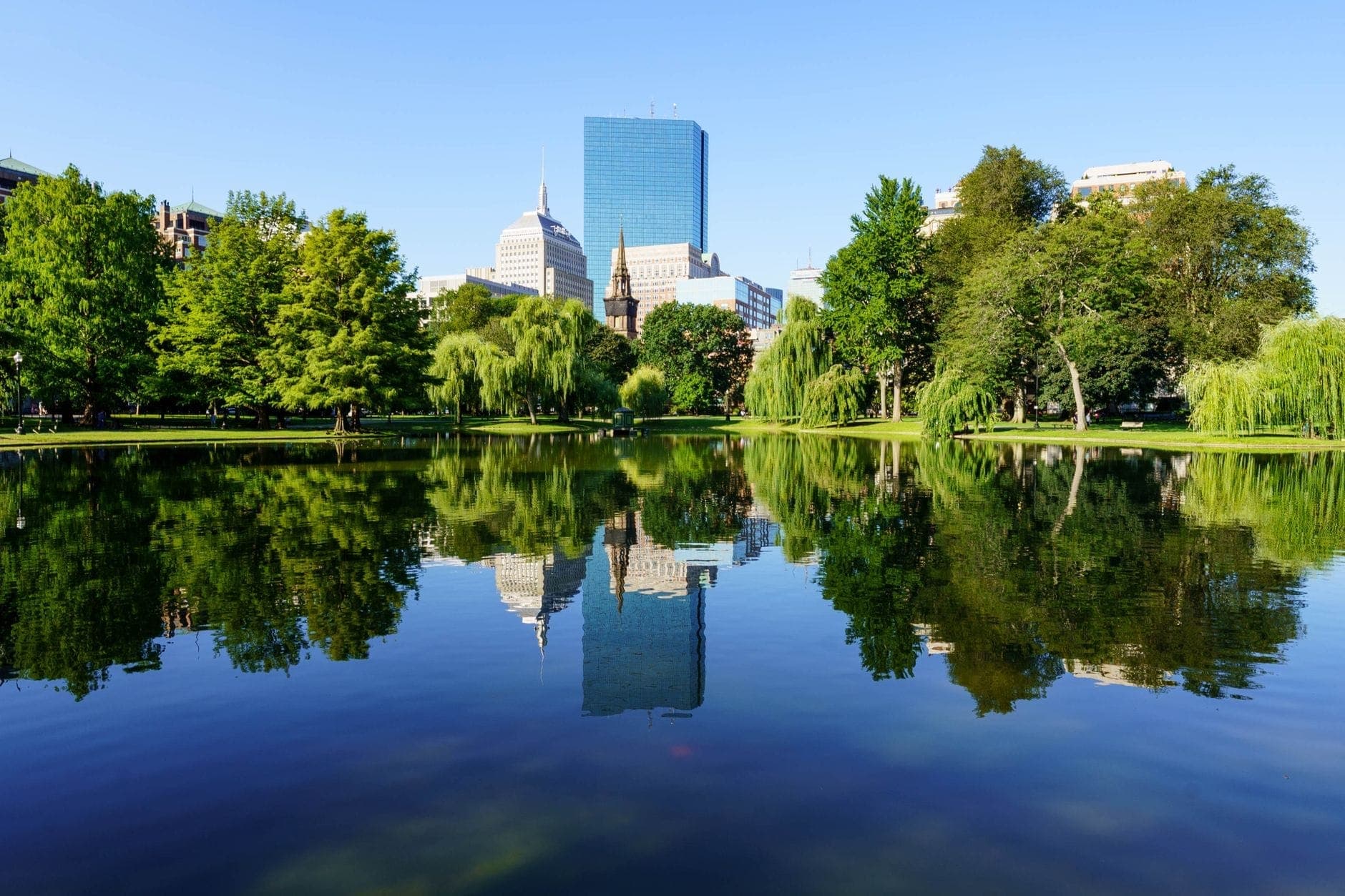 Skyline de Boston avec ses grands immeubles se reflétant dans les eaux calmes du Boston Public Garden par une journée ensoleillée, entouré d'arbres verdoyants.