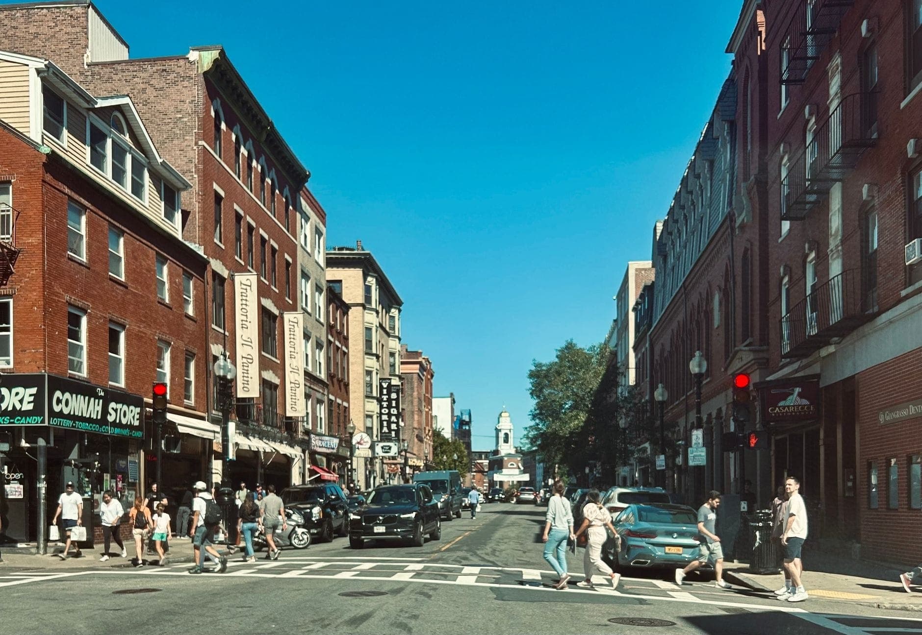 Rue animée dans le North End de Boston avec des bâtiments historiques en briques, des boutiques et des personnes traversant le carrefour par une journée ensoleillée.