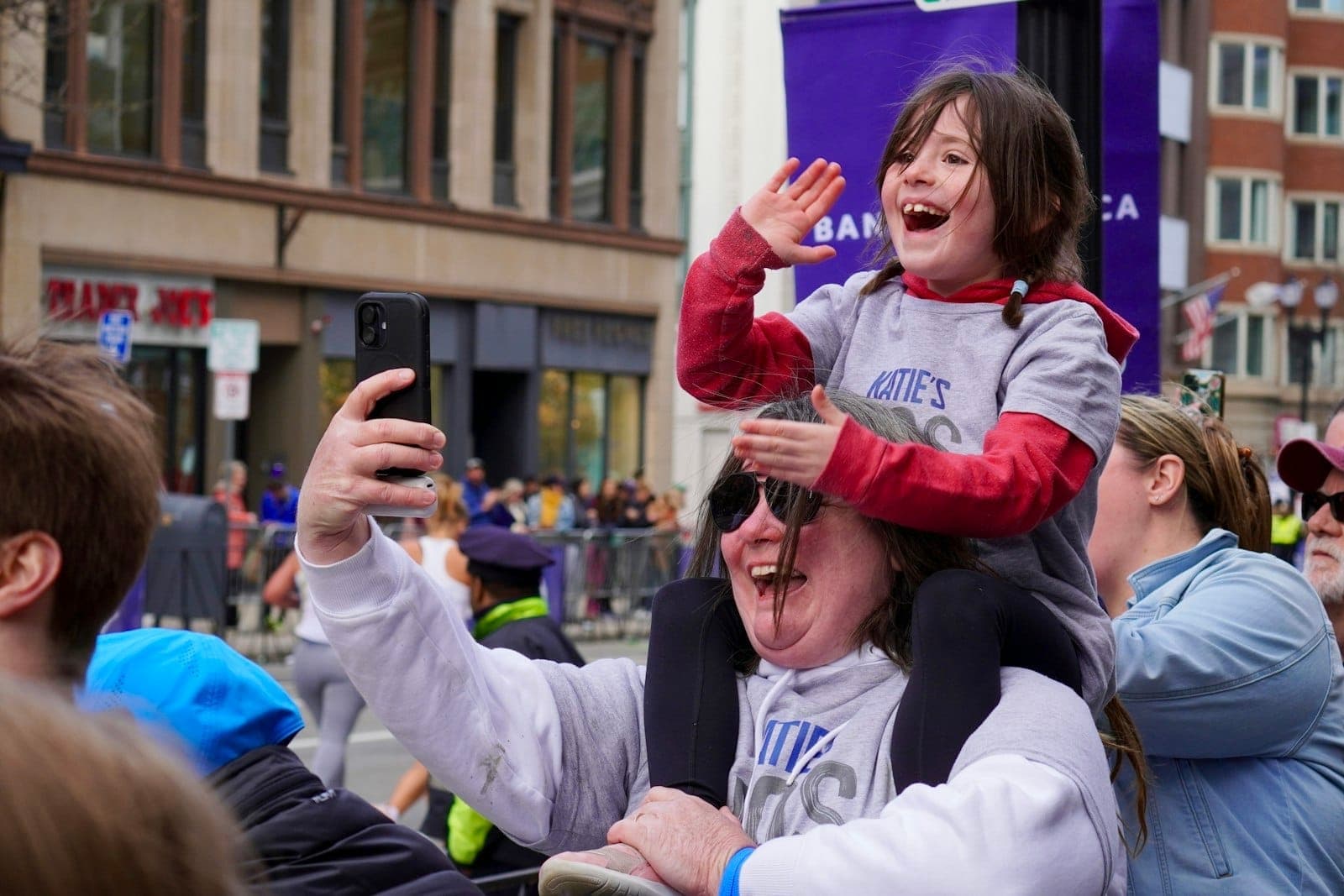 Un enfant souriant sur les épaules d'un adulte fait signe lors d'un événement animé dans une rue ensoleillée de Boston bordée de bâtiments historiques.
