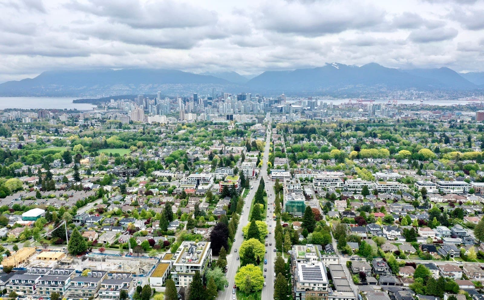 มุมมองทางอากาศของย่านสีเขียวและเส้นขอบฟ้าของเมือง Vancouver แสดงถนนที่เรียงรายด้วยต้นไม้และภูเขาในระยะไกลในวันที่มีเมฆมาก