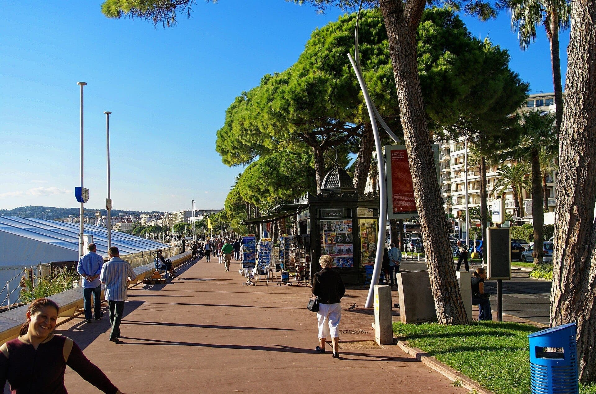 Люди прогуливаются вдоль усаженного пальмами Boulevard de la Croisette в Каннах, мимо газетных киосков и современных зданий под ясным голубым небом.