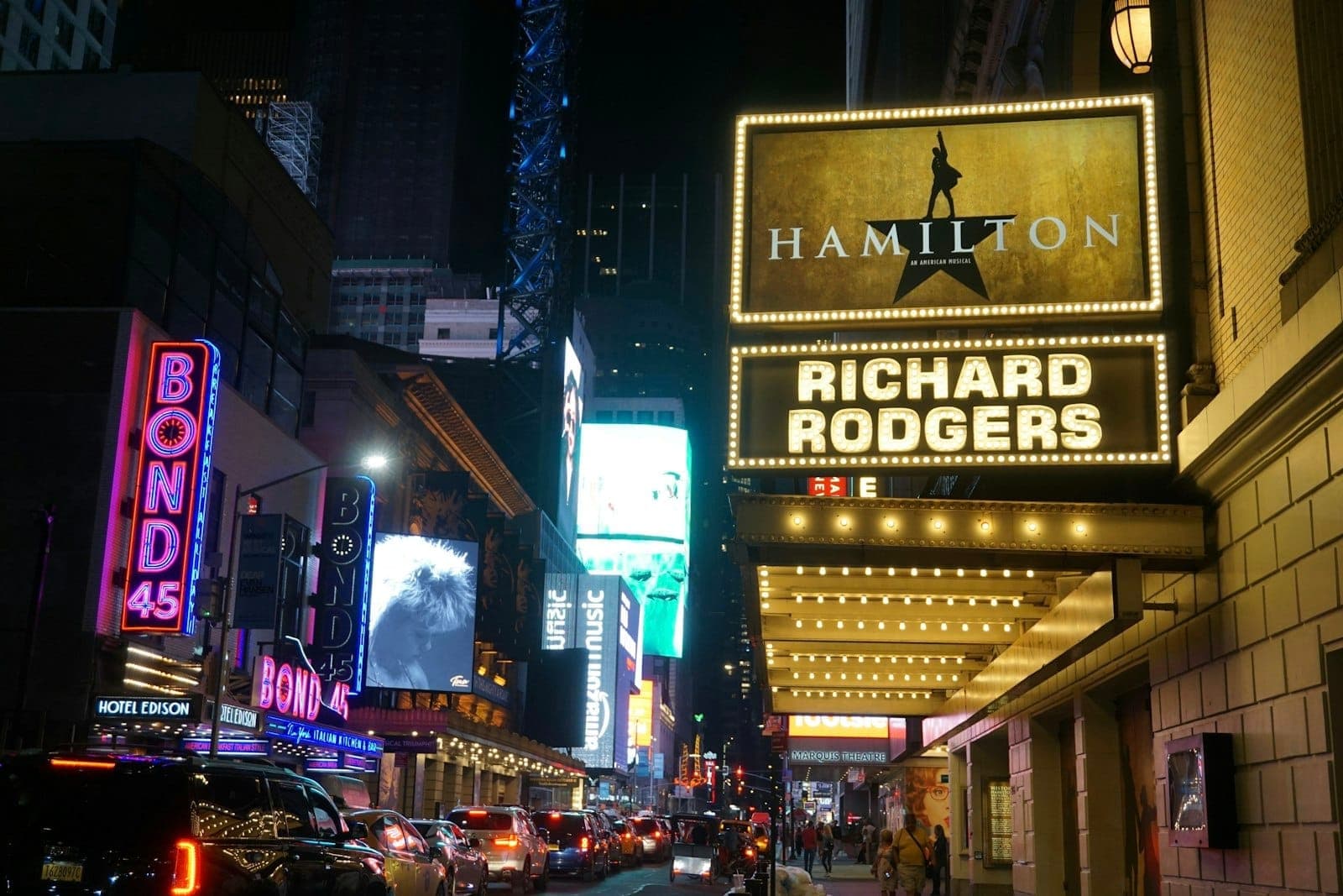 ภาพโรงละครบรอดเวย์เวลากลางคืน ป้ายไฟ Hamilton ที่ Richard Rodgers Theatre ผู้คนและรถหลายคันบนถนนนิวยอร์กที่คึกคัก