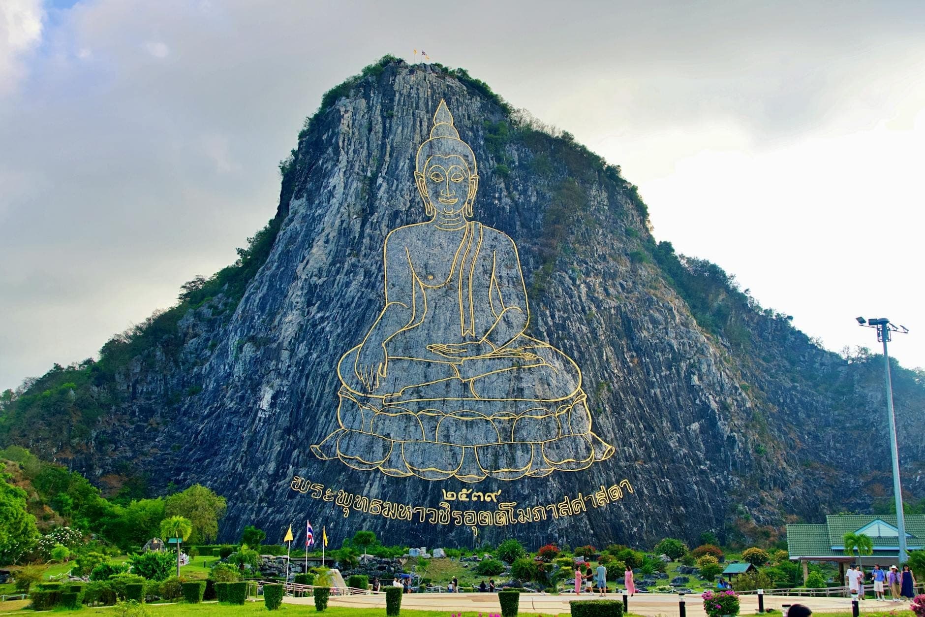 Огромный образ Будды, очерченный золотом и вырезанный лазером в известняковой горе Кхао Чи Чан, в окружении ухоженных садов; на переднем плане — несколько посетителей.