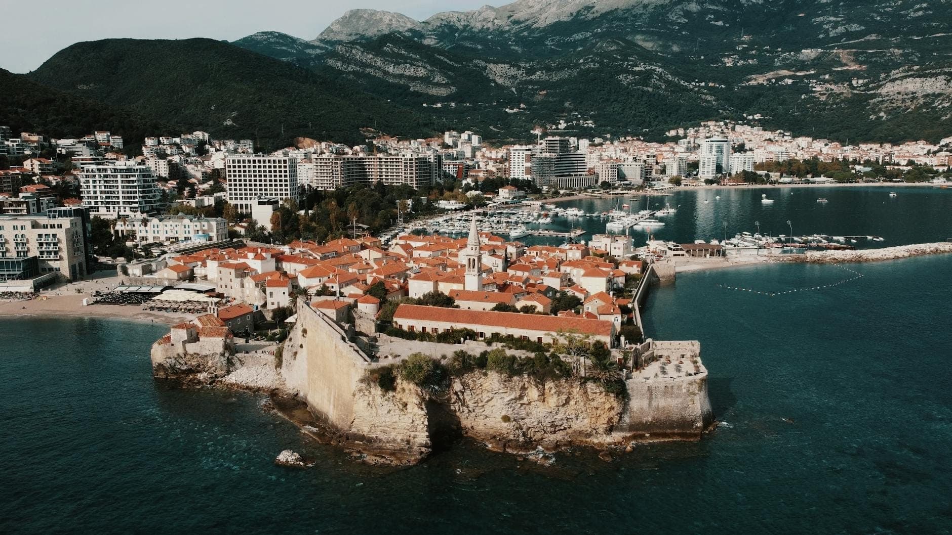Vue aérienne de la vieille ville et de la Citadelle de Budva sur la côte adriatique du Monténégro, avec ses remparts en pierre médiévaux, ses toits de tuiles rouges, la mer bleue et les montagnes en arrière-plan.