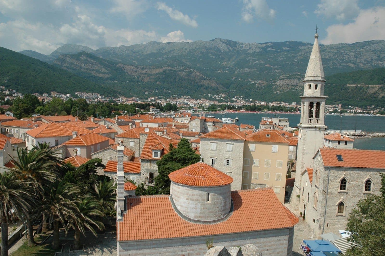 Vue sur la vieille ville médiévale de Budva avec ses toits de tuiles rouges, un clocher bien visible, les remparts vénitiens, les montagnes en arrière-plan et la mer Adriatique.