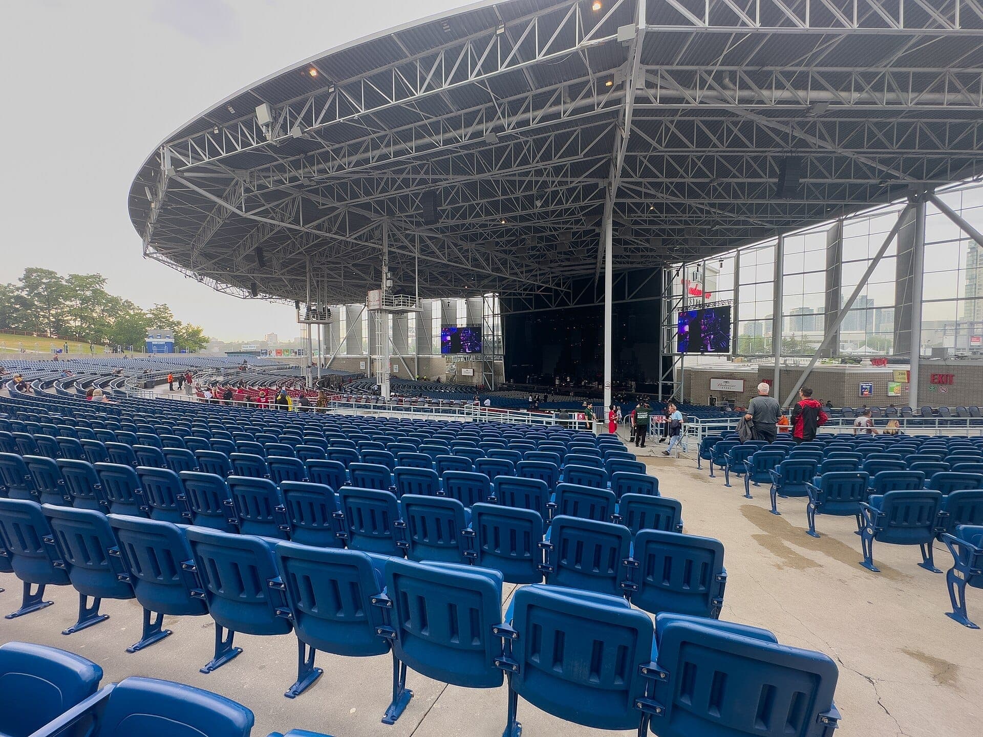 Vista panoramica del RBC Amphitheatre con le sue tribune a gradoni di colore blu sotto una moderna tettoia in acciaio, con alcune persone che si radunano prima di un concerto sul lungomare.