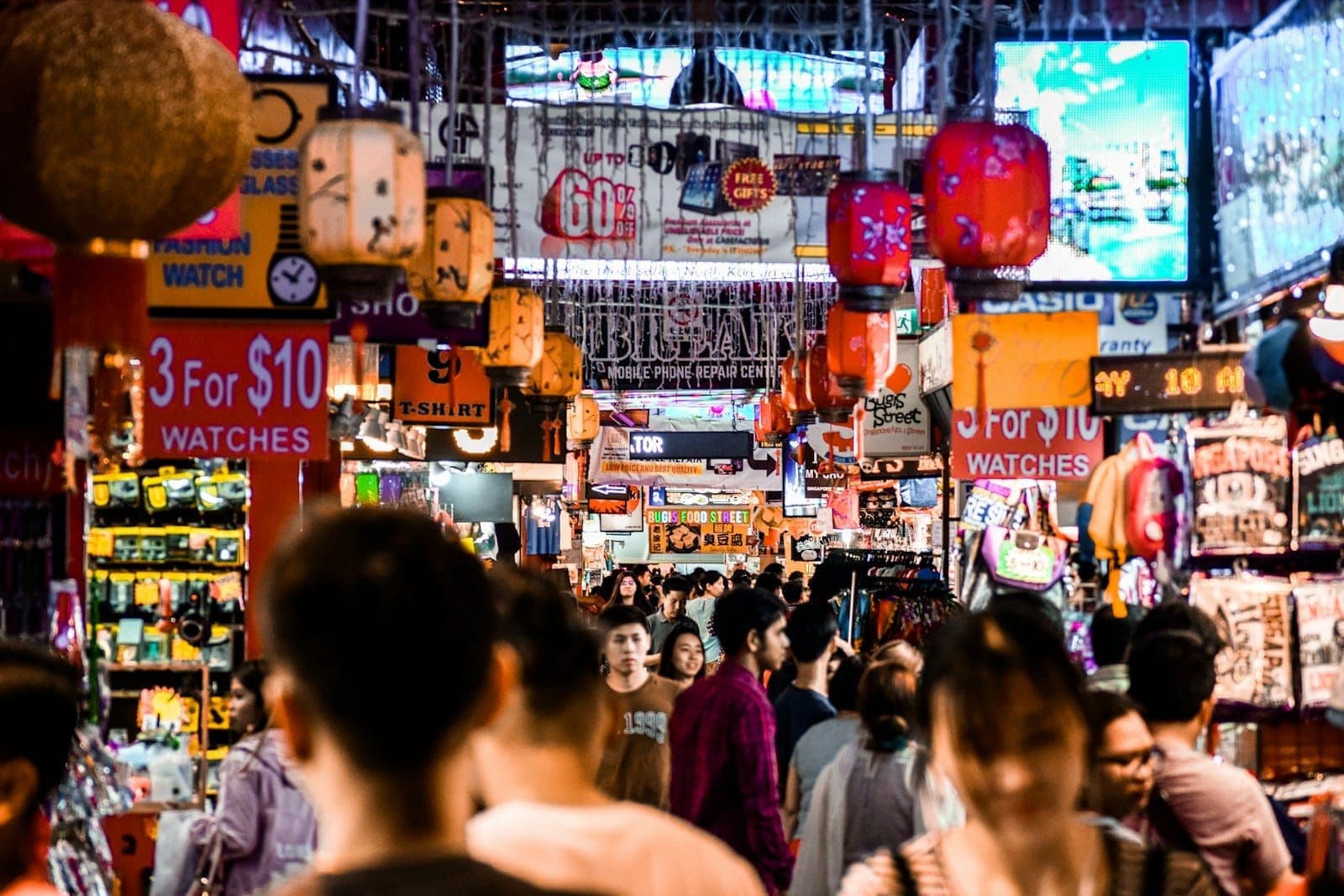 ทางเดินที่แออัดภายในตลาด Bugis Street Market พร้อมโคมไฟหลากสีสัน ป้ายสีสดใส และนักช้อปที่พลุกพล่านเดินเลือกของตามแผงขายของที่อัดแน่นใต้แสงไฟสว่างจ้า