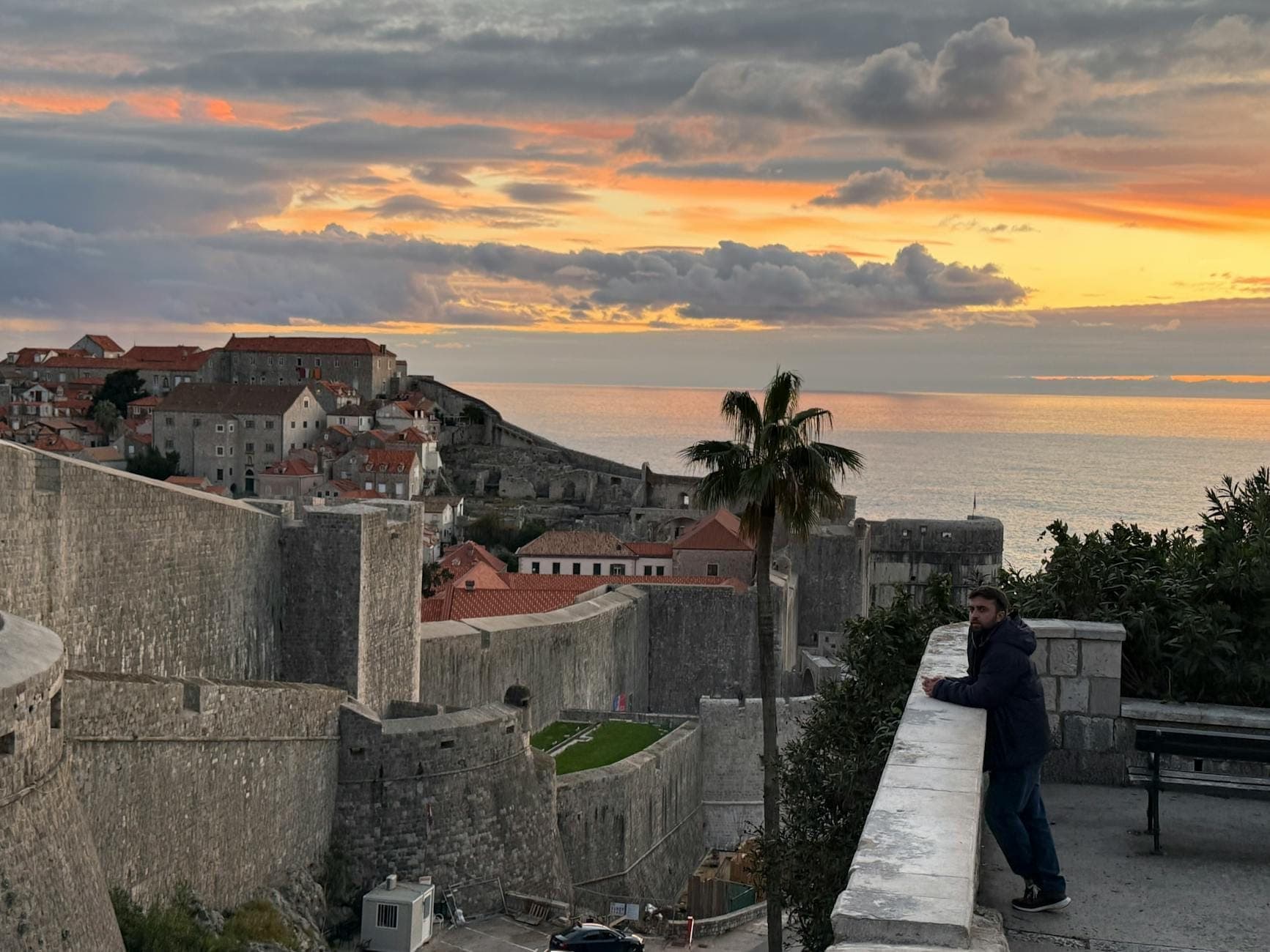 Vista de las murallas antiguas de Dubrovnik al atardecer, con el sol parcialmente ocultado por la muralla y un hombre mirando el mar.