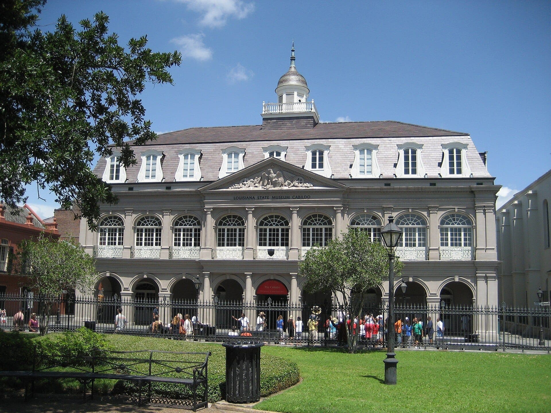 Le Cabildo à La Nouvelle-Orléans, un grand bâtiment historique avec des fenêtres en arc et un dôme, se dresse derrière une clôture en fer forgé et des pelouses soignées par une journée ensoleillée.