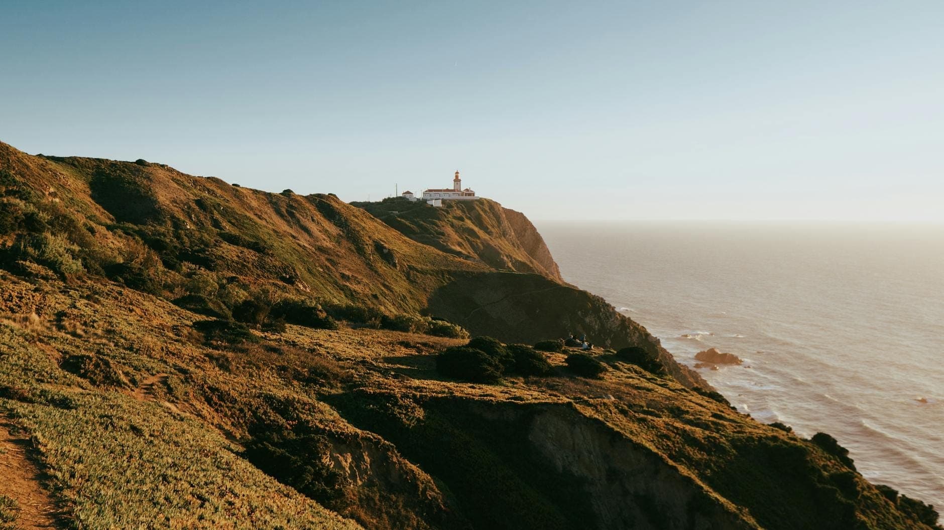 Vista ampla das falécias imponentes do Cabo da Roca se erguendo acima do Oceano Atlântico, com o farol posicionado na beira do precipício sob um céu limpo.