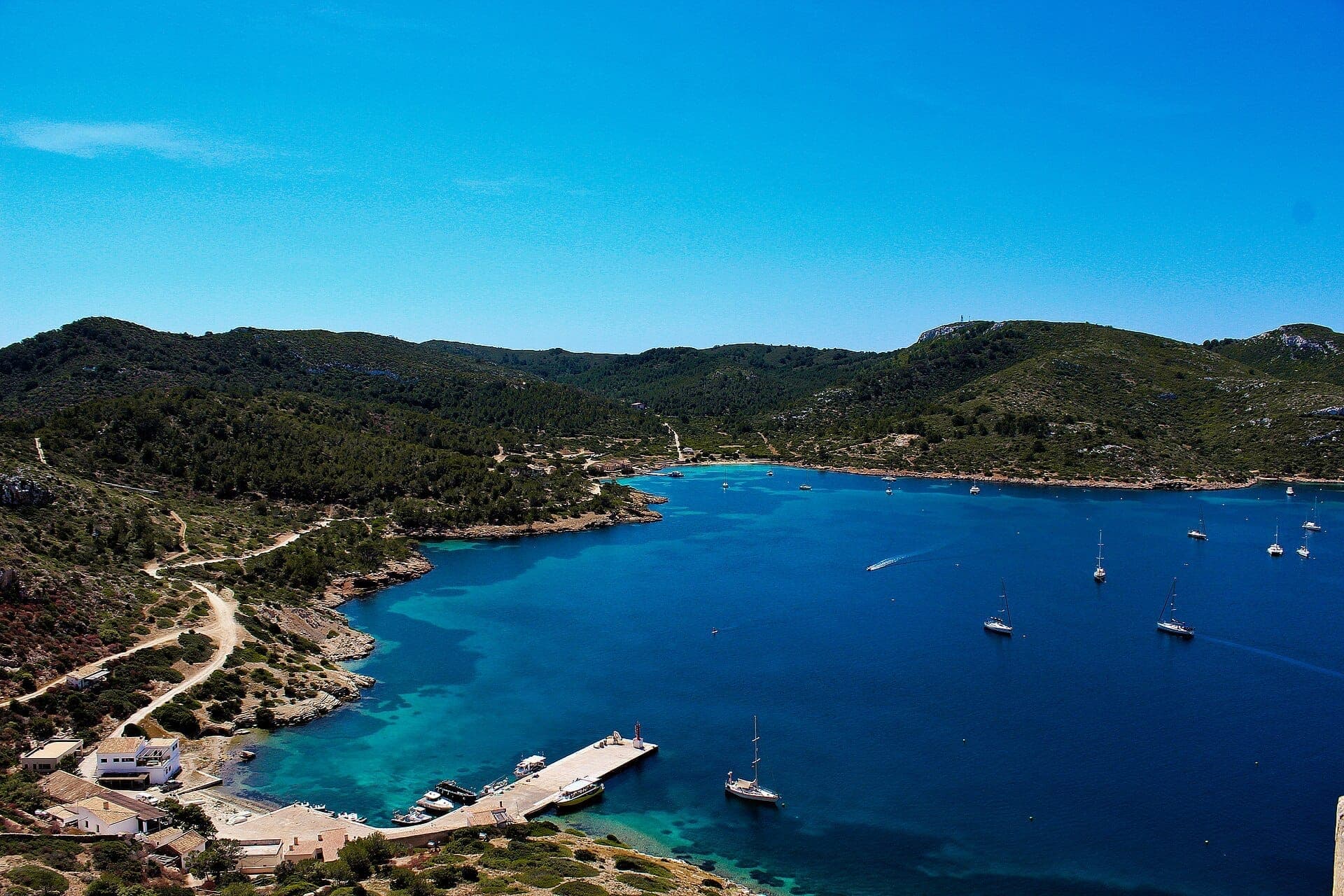 Luftaufnahme des Nationalparks Cabrera mit einer kristallklaren blauen Bucht, bewaldeten Hügeln, einem kleinen Yachthafen und vereinzelten Segelbooten.