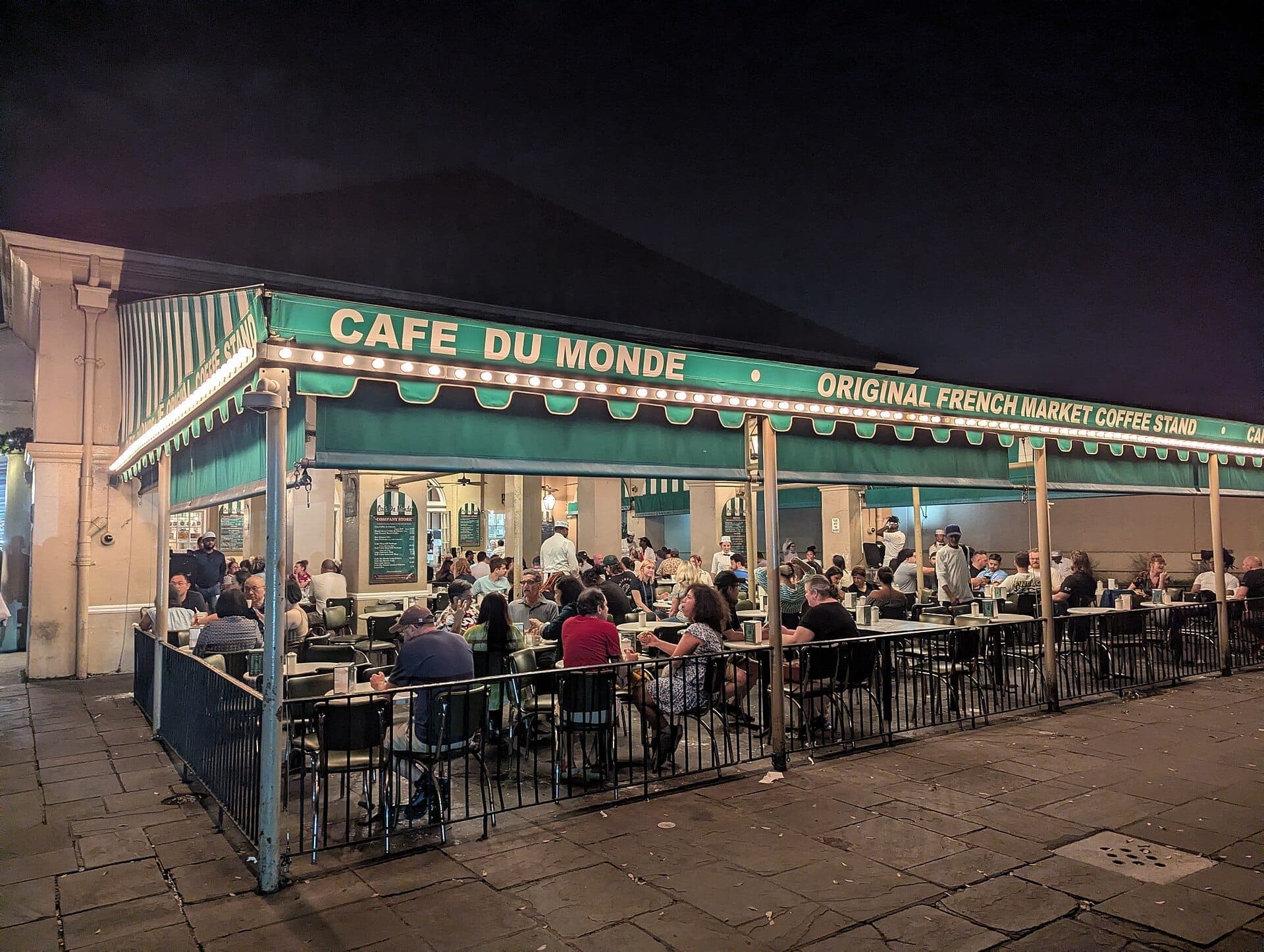 Открытая терраса Café du Monde ночью: люди наслаждаются бенье и кофе под зелёным навесом в окружении тёплых гирляндных огней.
