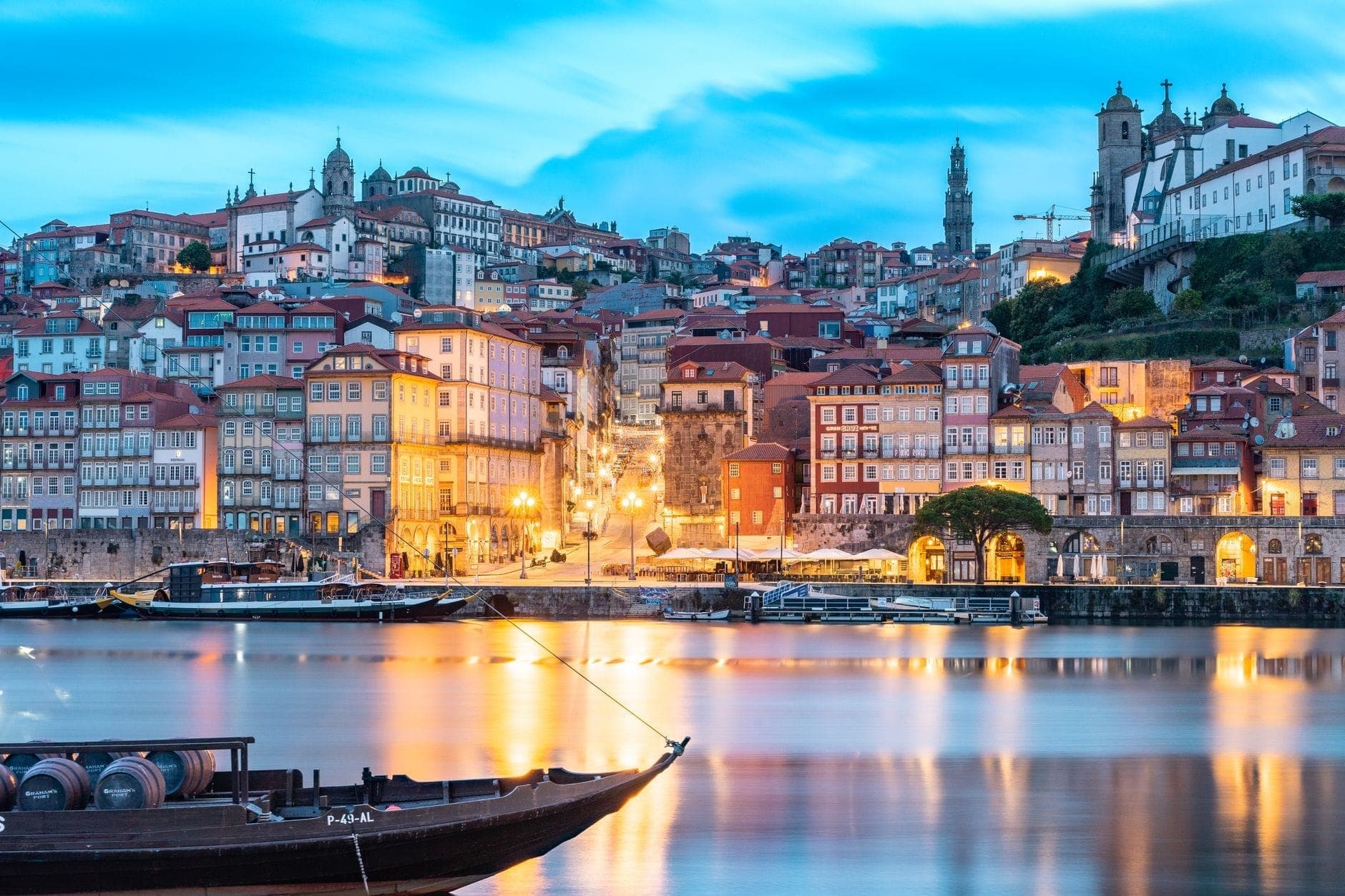 Le Cais da Ribeira de Porto illuminé au crépuscule, avec ses bâtiments historiques colorés, sa promenade au bord du fleuve et ses bateaux traditionnels reflétés dans le Douro sous un ciel bleu intense.