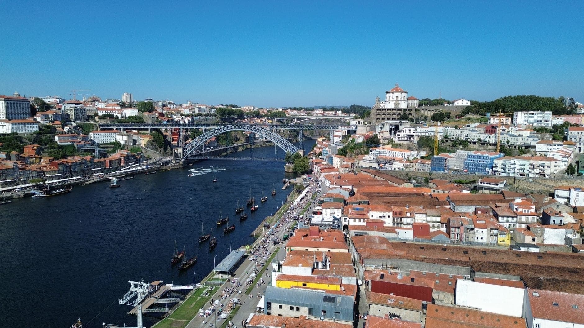 Luftaufnahme des Cais de Gaia mit Rabelo-Booten auf dem Douro, der Dom-Luís-I-Brücke und Portos bunter Skyline unter strahlend blauem Himmel.