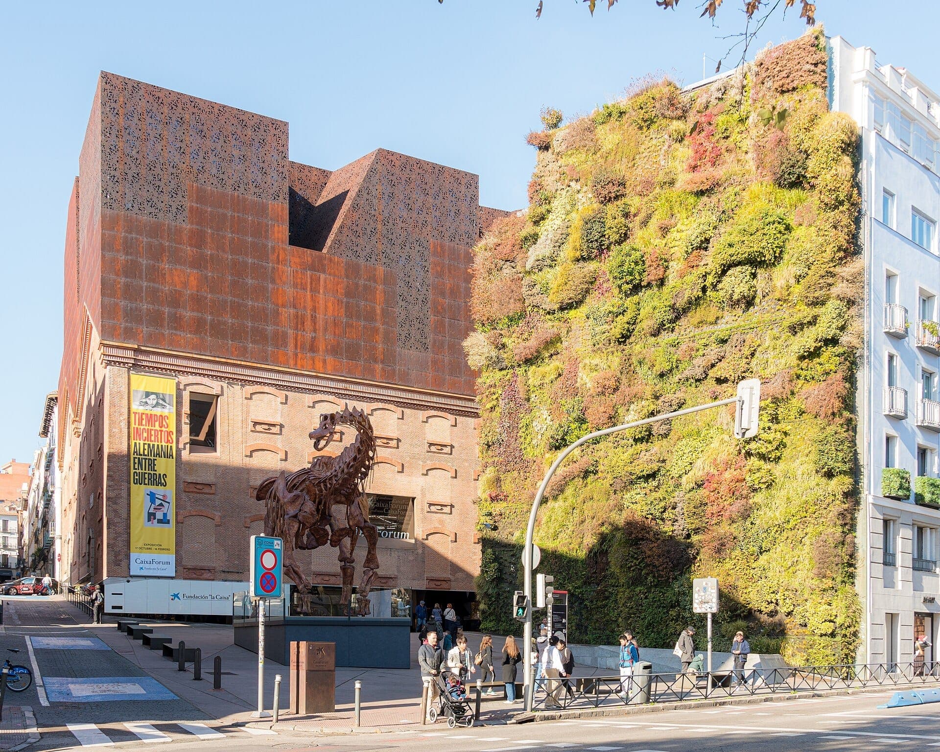 ภายนอก CaixaForum Madrid ที่มีสถาปัตยกรรมสีสนิมอันเป็นเอกลักษณ์ สวนแนวตั้งขนาดใหญ่ ผู้คนกำลังเดิน และประติมากรรมม้าที่โดดเด่นใต้ท้องฟ้าสีครามแจ่มใส