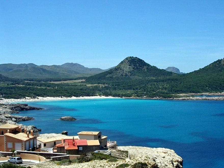 Blick auf den Strand Cala Agulla auf Mallorca mit türkisblauem Wasser, Kiefernwald im Hintergrund, Hügeln und einigen Häusern entlang der felsigen Küste.