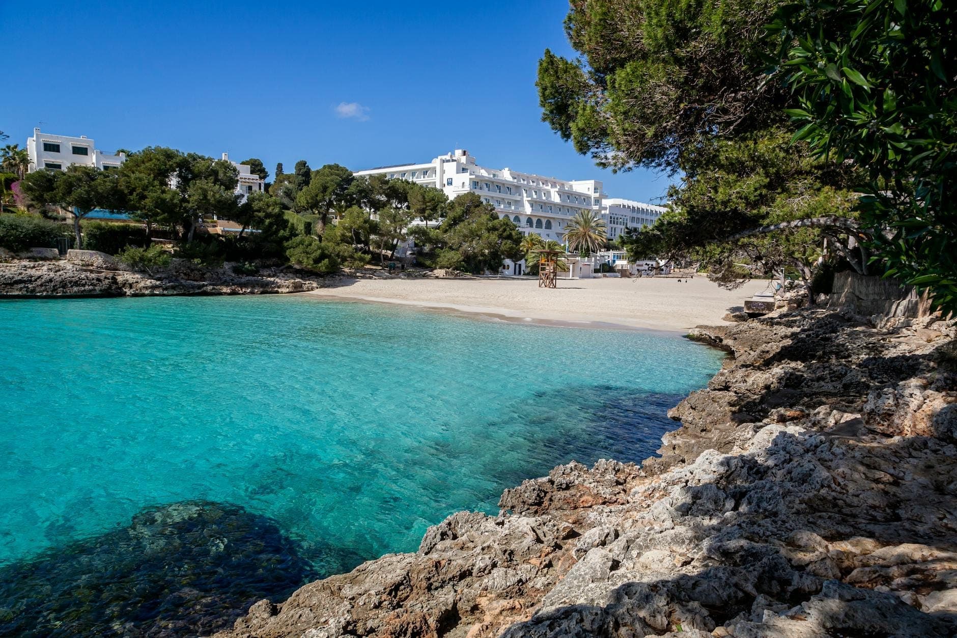 Bucht mit türkisfarbenem Wasser, weißem Sandstrand und felsigem Ufer im Vordergrund; weißgetünchte Gebäude und Bäume im Hintergrund unter klarem blauem Himmel.