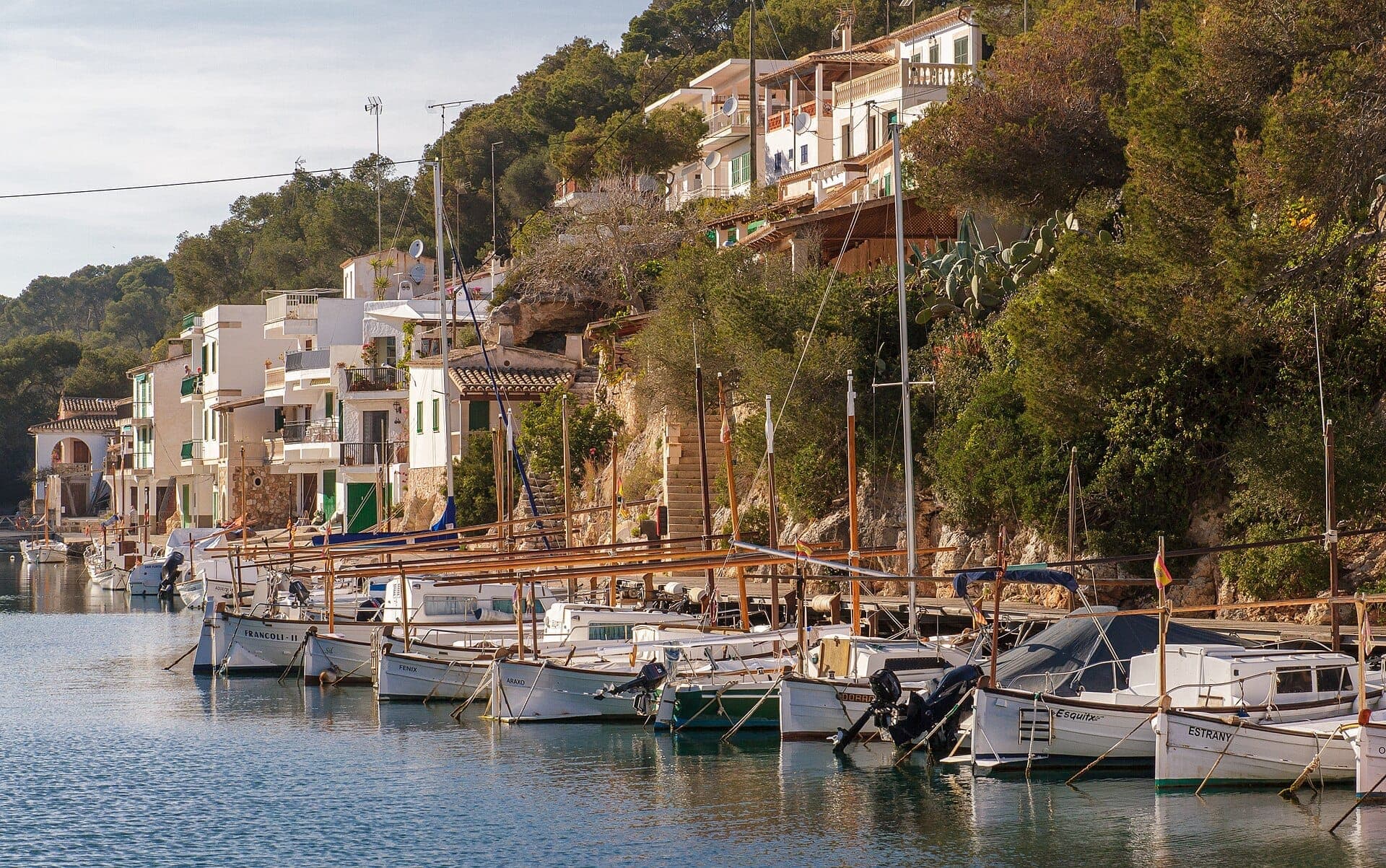 Traditionelle Fischerboote liegen an den ruhigen Gewässern von Cala Figuera vor Anker, mit weiß getünchten Häusern und üppigem Grün, das den Hang hinaufklettert