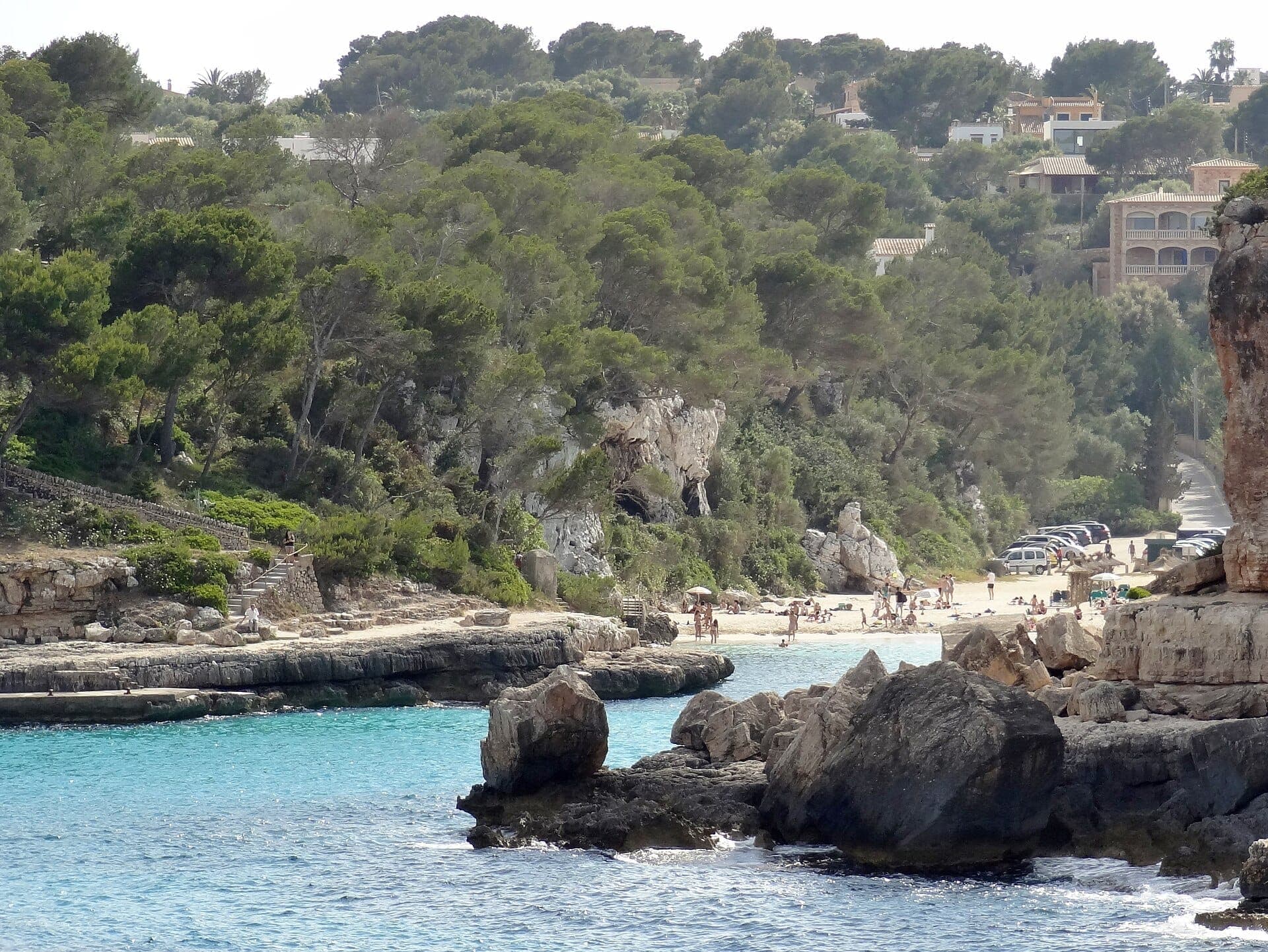 Strand von Cala Llombards mit türkisfarbenem Wasser, weißem Sand, Kalksteinfelsen, vereinzelten Sonnenbadenden, Pinienbäumen und Hangvillen im Hintergrund.