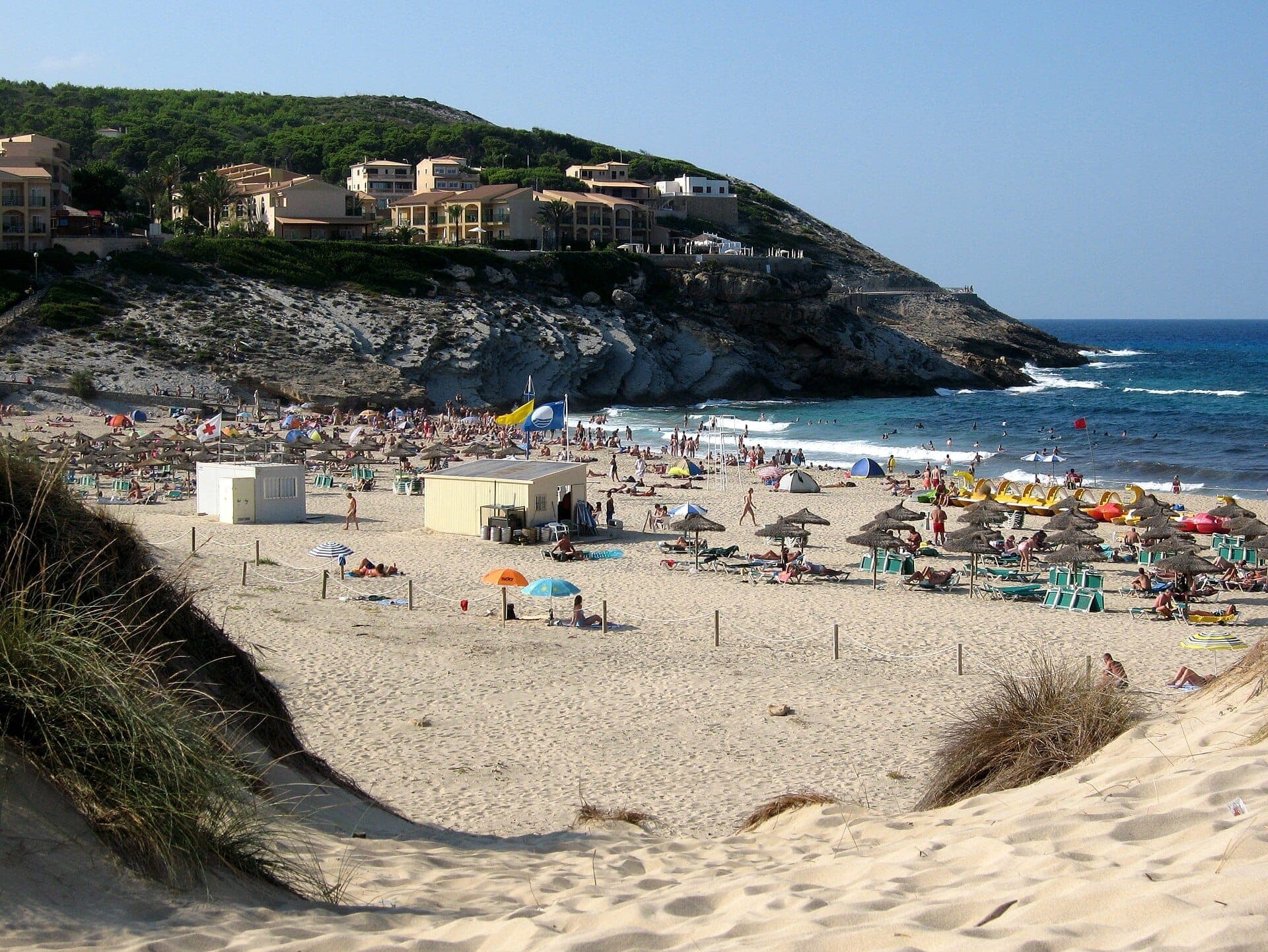Strand Cala Mesquida mit goldenen Sanddünen, Liegestühlen, Sonnenschirmen und türkisfarbenen Wellen, eingerahmt von Felsenklippen und Hanggebäuden.