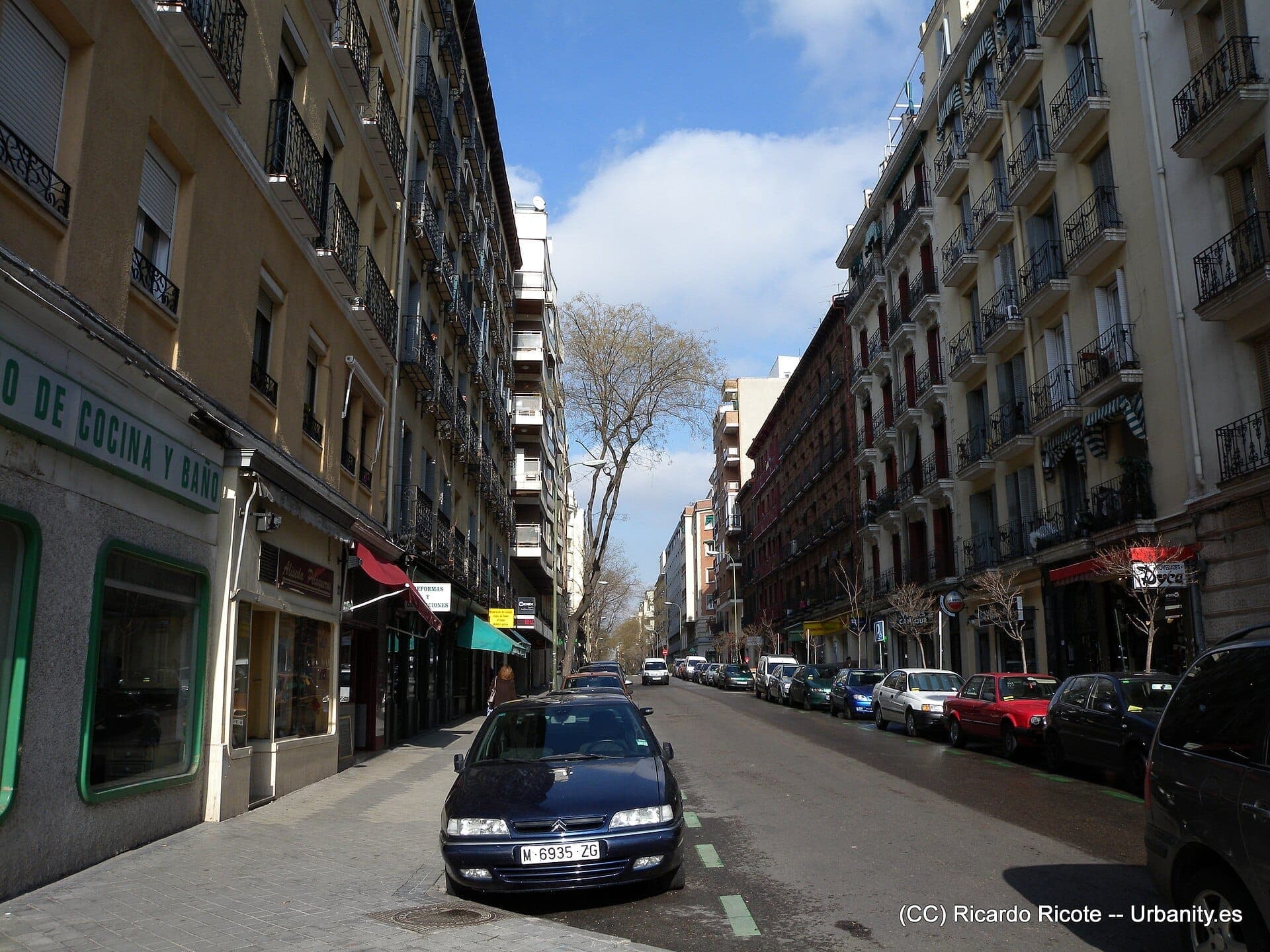 วิวถนน Calle de Ponzano ในมาดริด ซึ่งมีอาคารอพาร์ตเมนต์สูงใหญ่ รถยนต์จอดเรียงราย และป้ายร้านค้าในวันที่แดดจัด