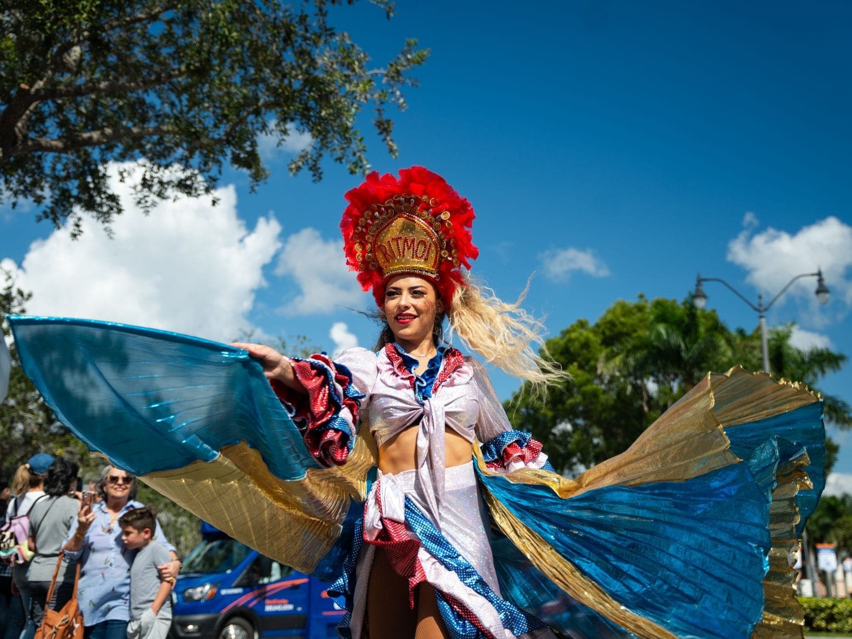 Woman in elaborate, colorful carnival costume with feathered headdress dances outdoors during a street festival under a bright blue sky with onlookers nearby.