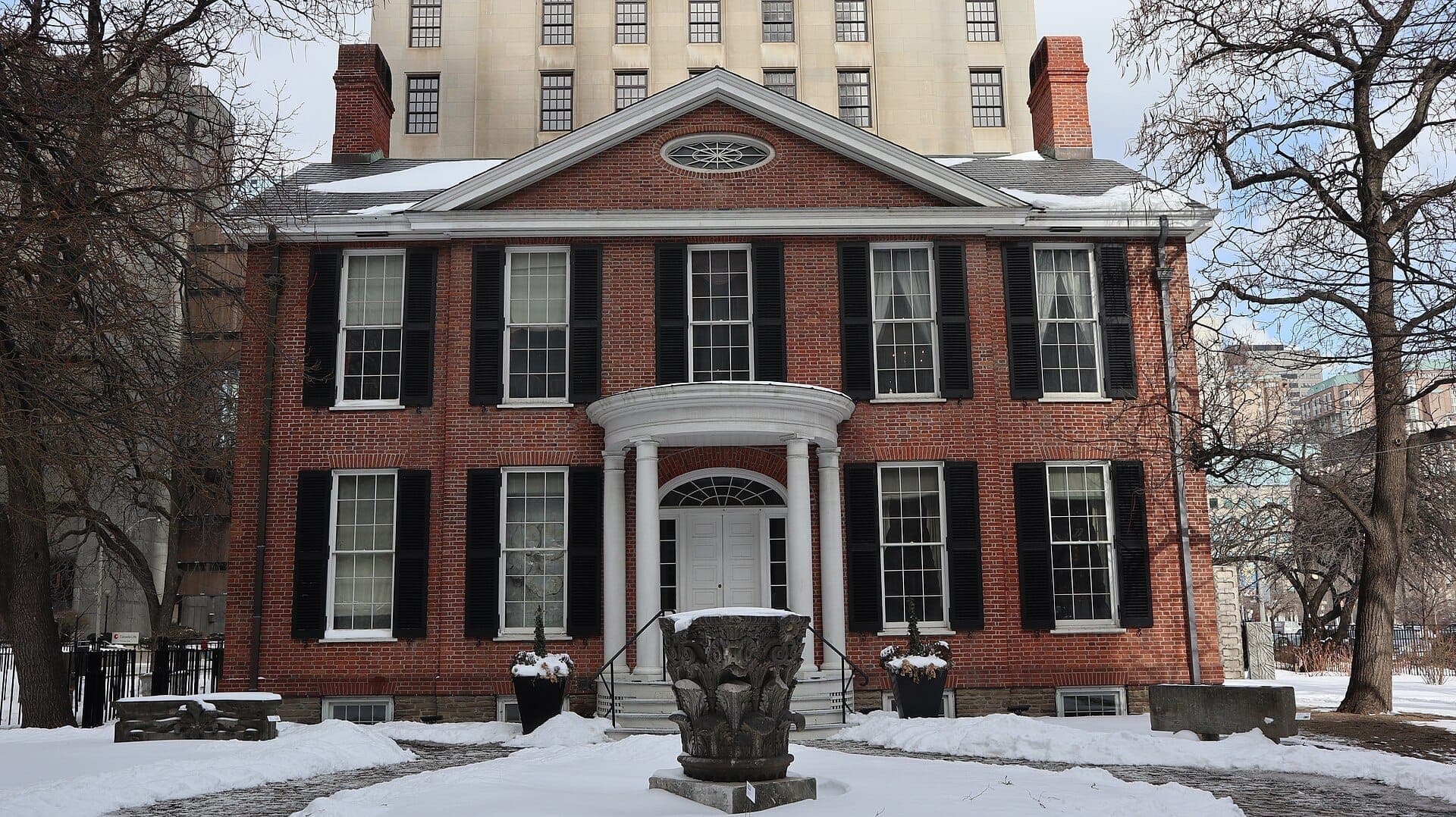 Il Campbell House Museum, un edificio georgiano in mattoni rossi con persiane nere, si erge nella neve con una grande struttura del centro città sullo sfondo.