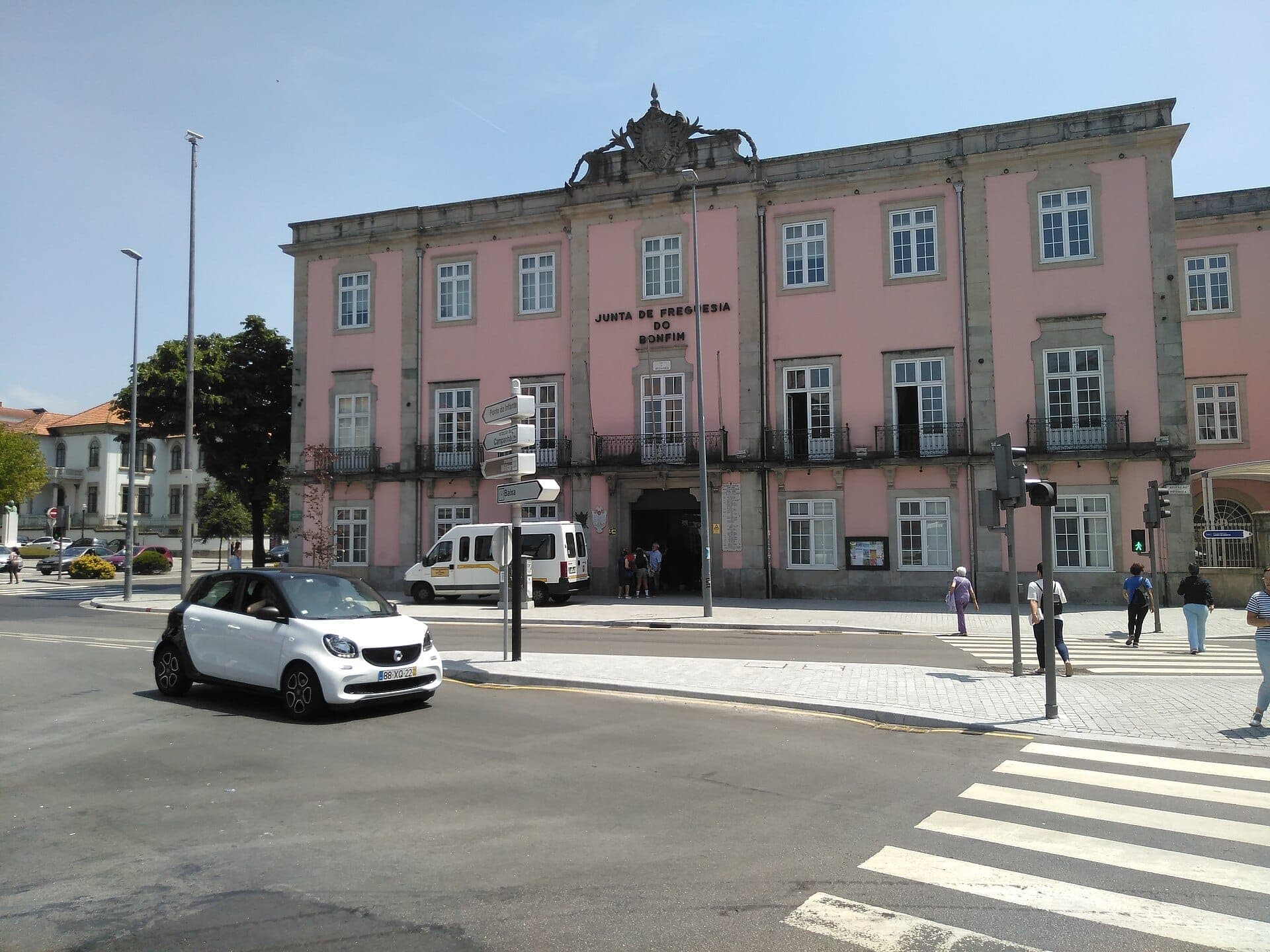Edificio storico rosa in piazza Campo 24 de Agosto a Porto, con persone che attraversano la strada e auto che passano sotto un cielo azzurro limpido.