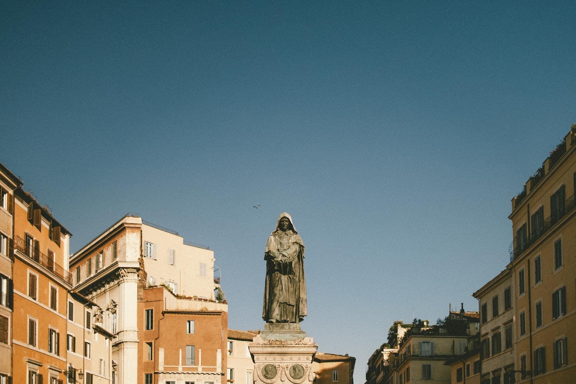 Rebulto ni Giordano Bruno sa gitna ng Campo de' Fiori sa Roma, napapalibutan ng mga makasaysayang gusali at maliwanag na asul na langit.