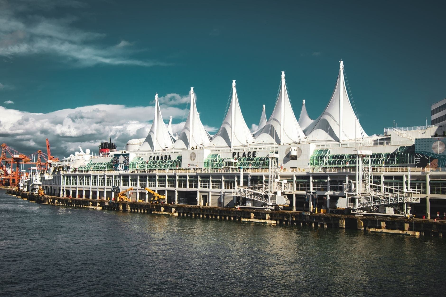 ภาพกว้างของหลังคาทรงใบเรือสีขาวอันเป็นสัญลักษณ์ของ Canada Place ริมน้ำแวนคูเวอร์ ข้างอ่าวและท้องฟ้าดราม่าพร้อมเงาสะท้อนน้ำ เชิญชวนให้อยากมาเยือน