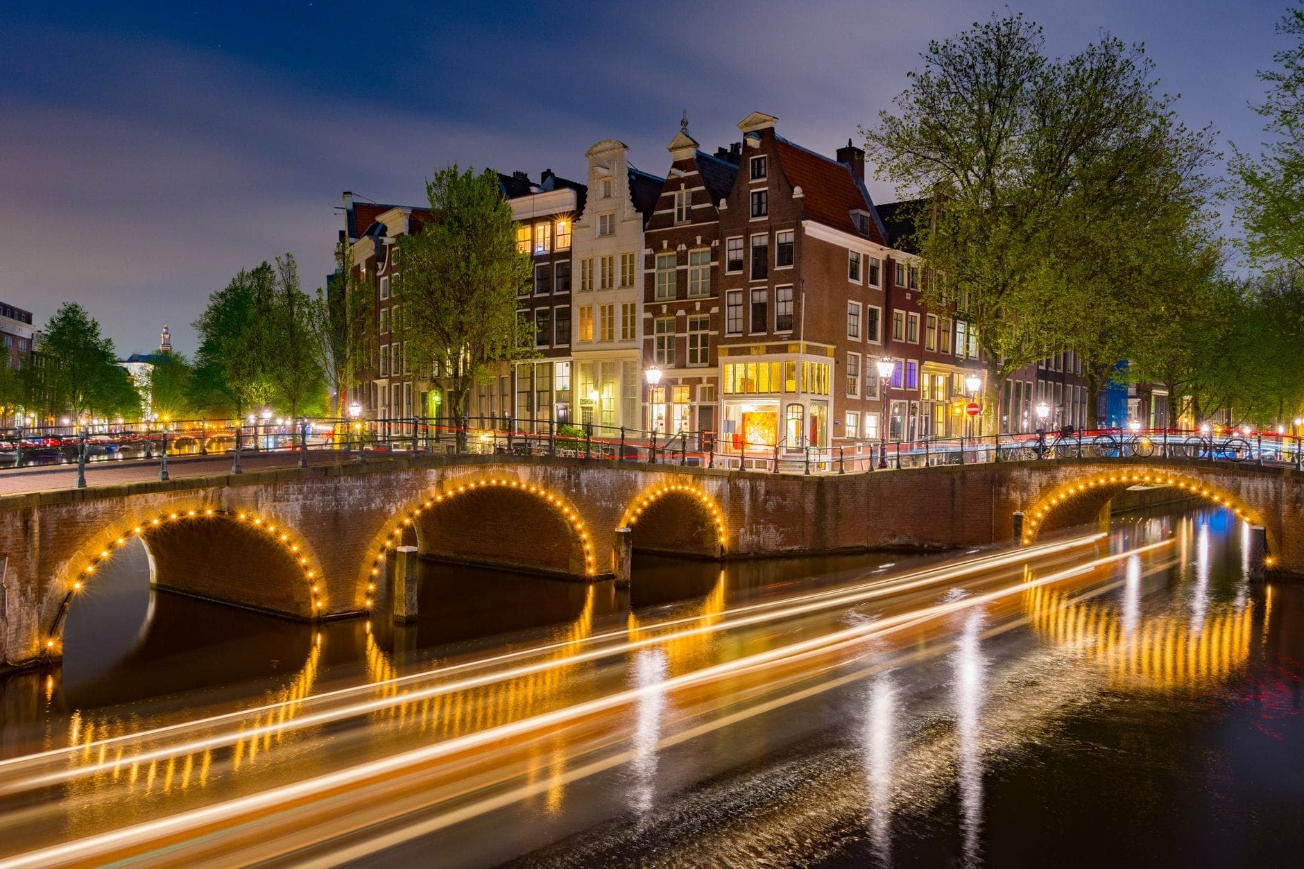 Nächtlicher Blick auf einen Amsterdamer Kanal mit beleuchteten Bogenbrücken, klassischen Kaufmannshäusern und leuchtenden Bootsspuren, die sich im Wasser spiegeln.
