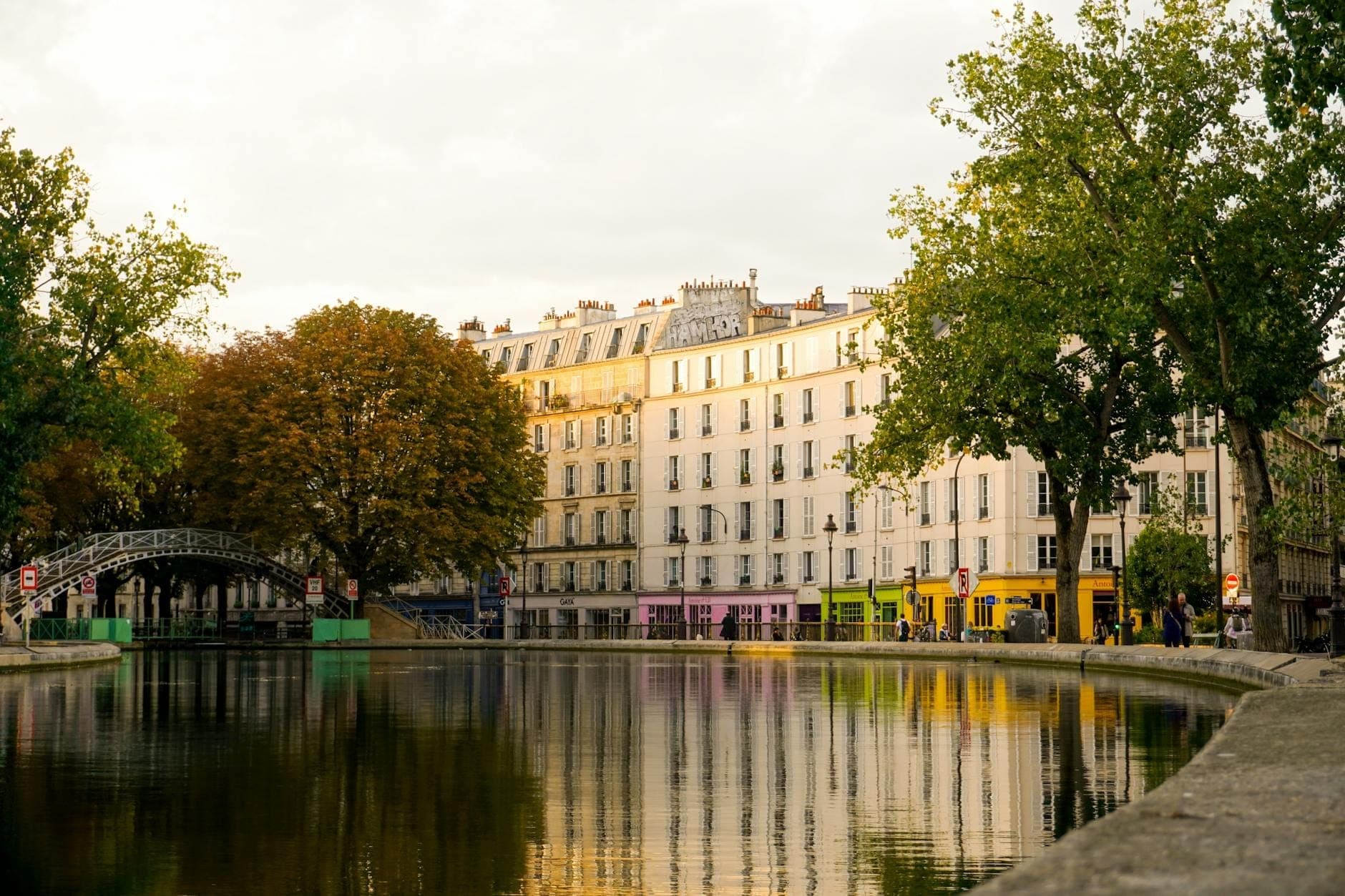 The Canal Saint-Martin with calm water reflecting colorful Parisian buildings and autumn trees, an iron footbridge crossing the canal, and a peaceful, cinematic atmosphere.