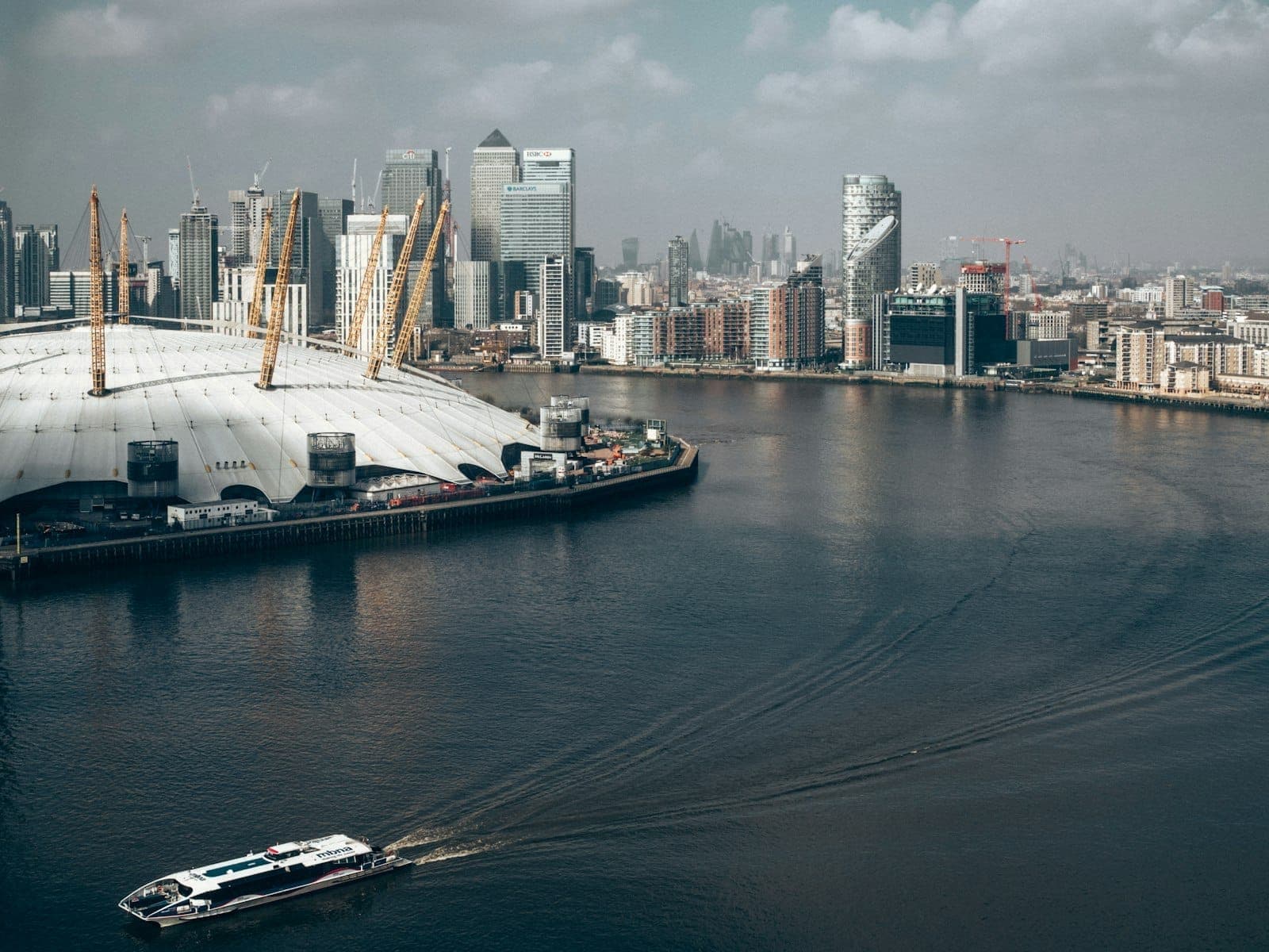 Vista do Tâmisa em direção à Arena O2 e ao skyline de Canary Wharf, com arranha-céus modernos, edifícios à beira do rio e um barco na água sob um céu nublado.