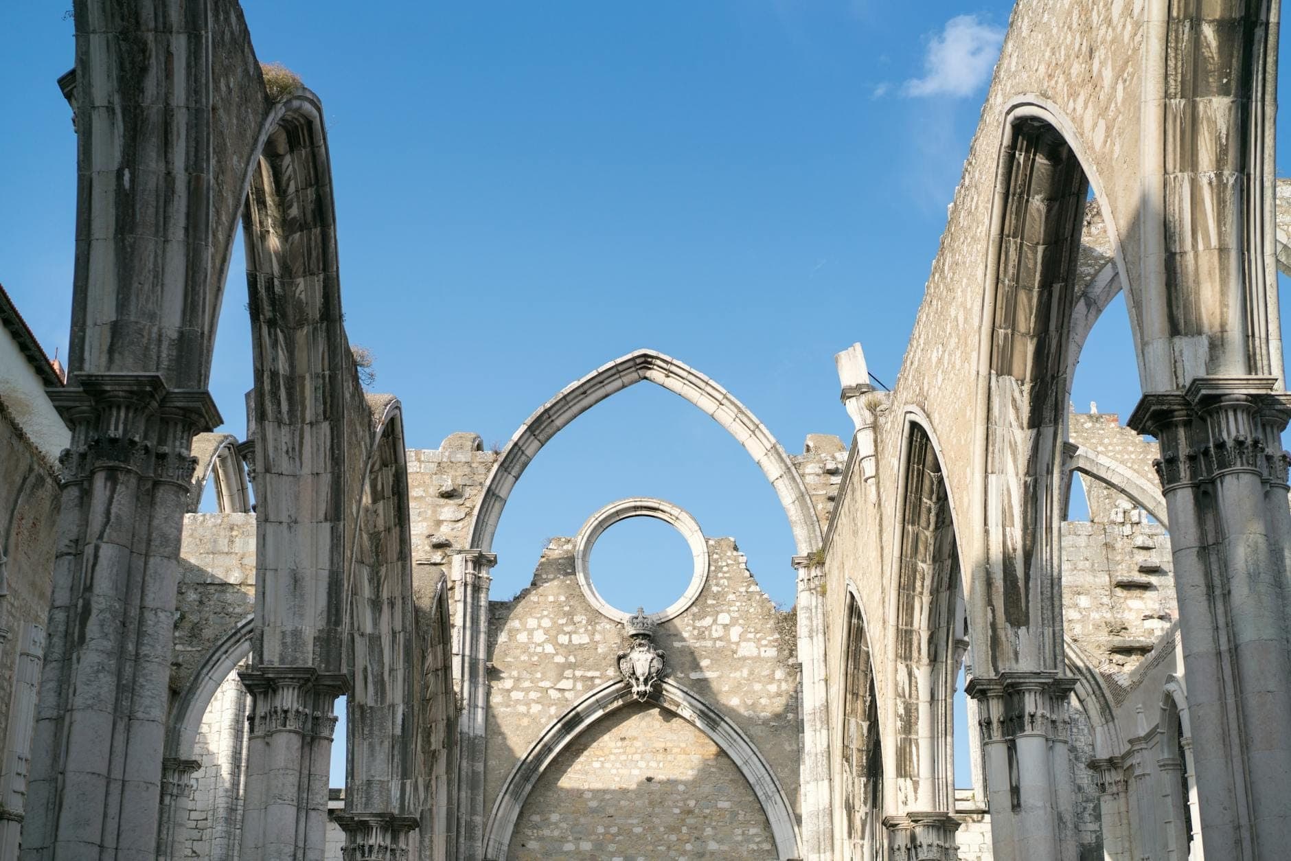 Arcos góticos em ruínas e paredes de pedra do Convento do Carmo em Lisboa, abertos para um céu azul intenso, transmitindo uma forte sensação de história e atmosfera.