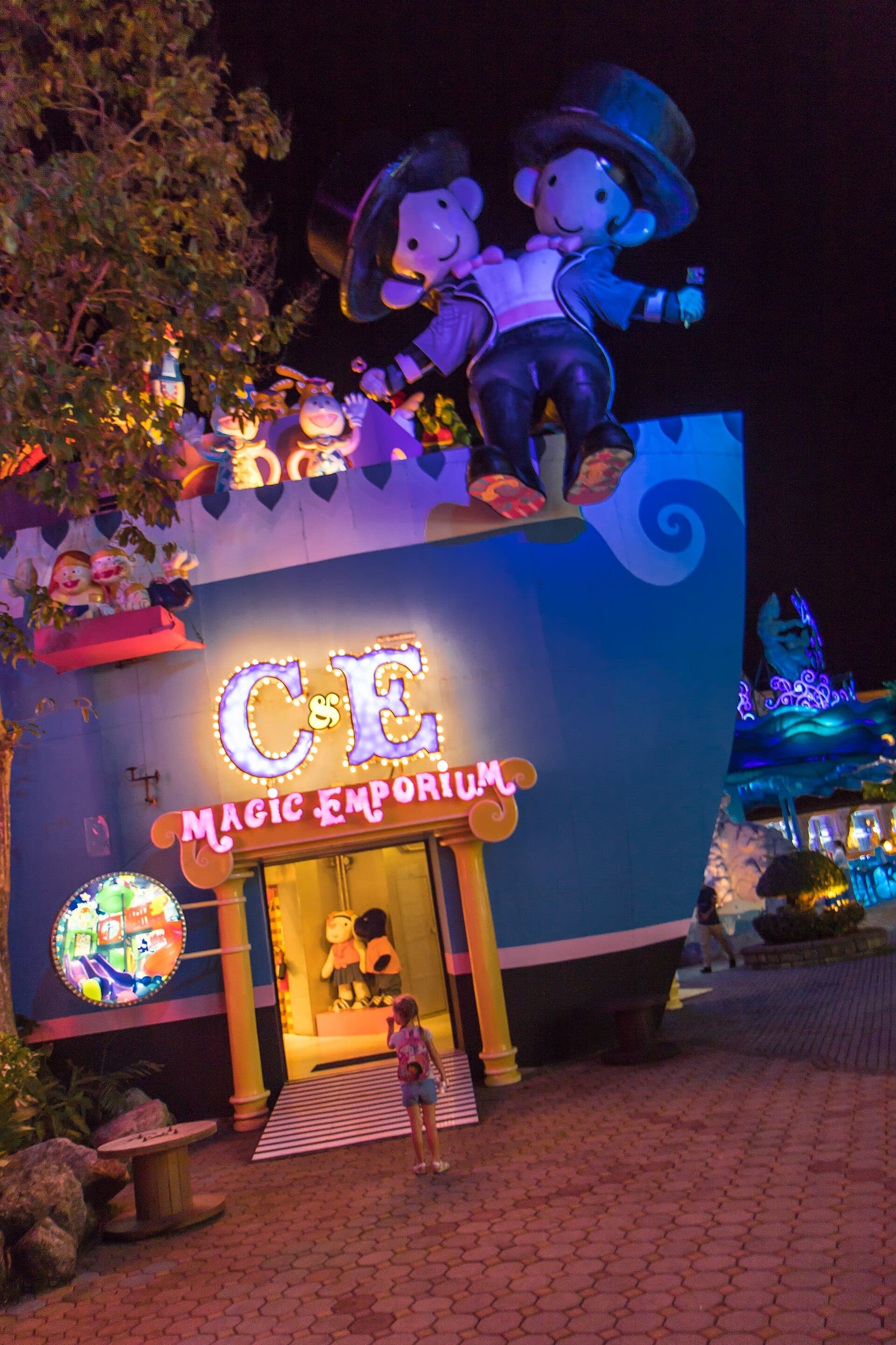 Carnival Magic Phuket's Magic Emporium at night, featuring colorful cartoon statues, neon lights, and a young visitor admiring the entrance.