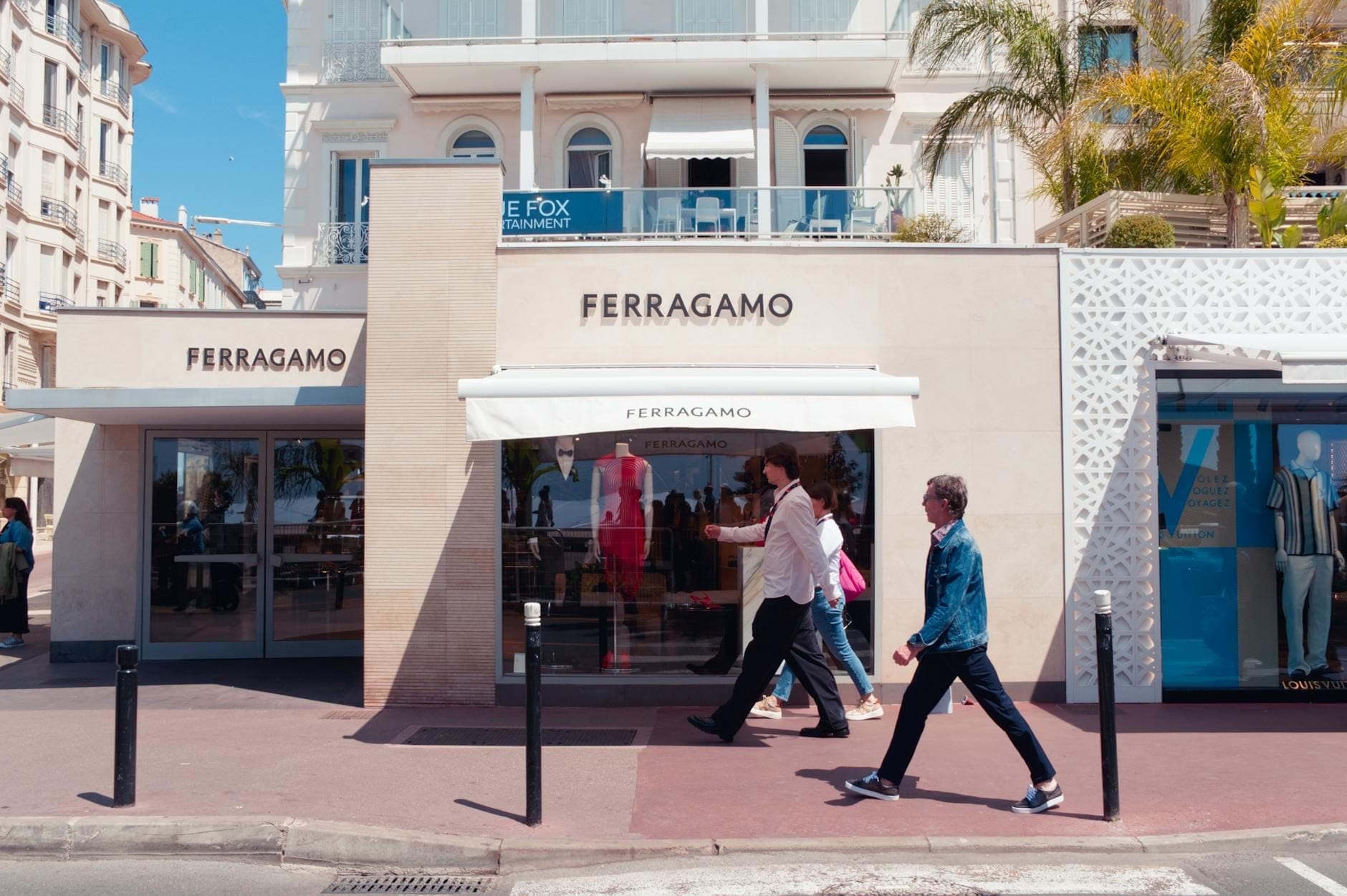 Фасад магазина Ferragamo в Carré d'Or в Каннах: стильные прохожие и пальмы, обрамляющие элегантные белые здания на заднем плане.