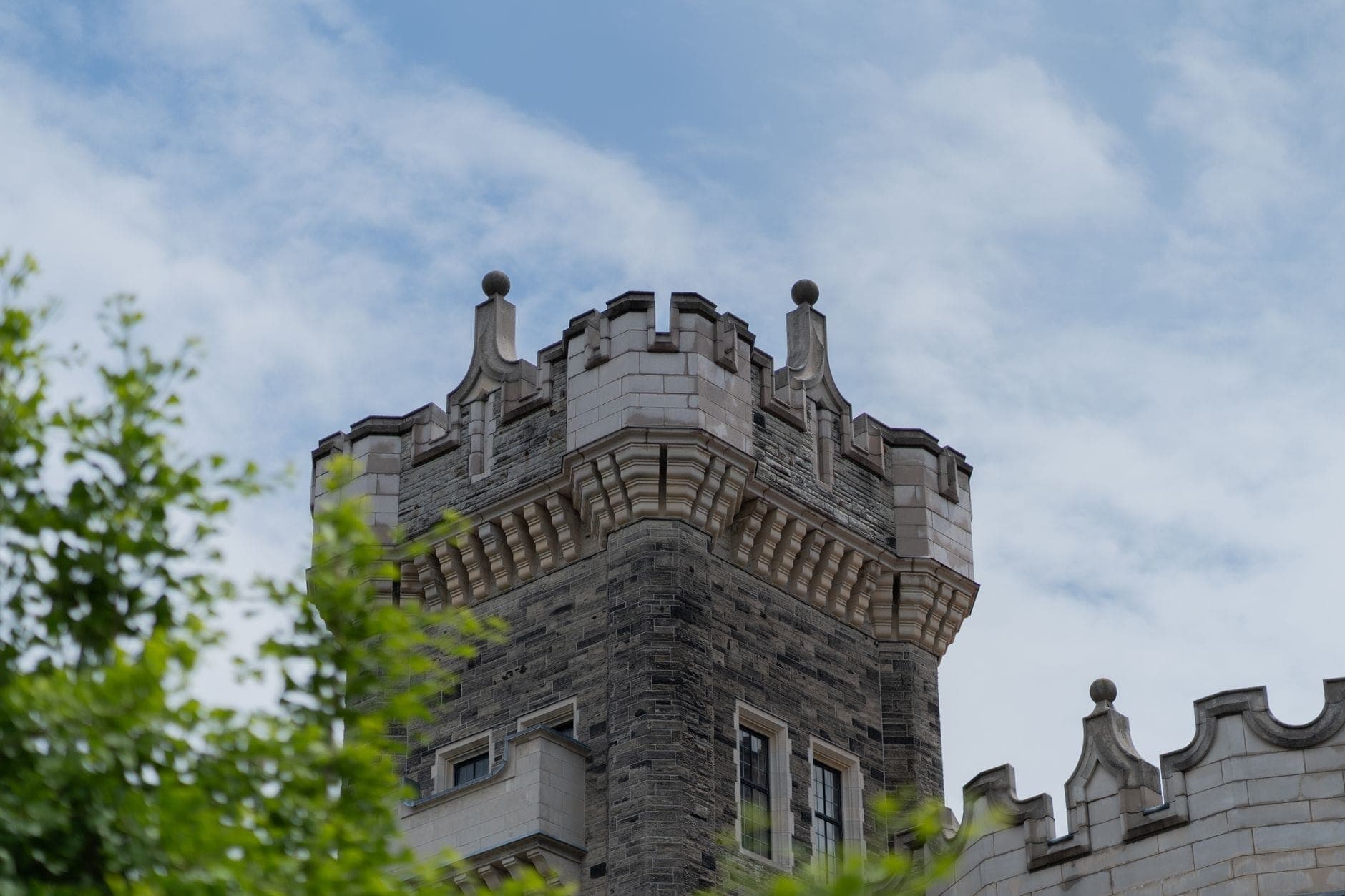 Vista ravvicinata della caratteristica torre merlata di Casa Loma, incorniciata da rami frondosi e un cielo parzialmente nuvoloso.