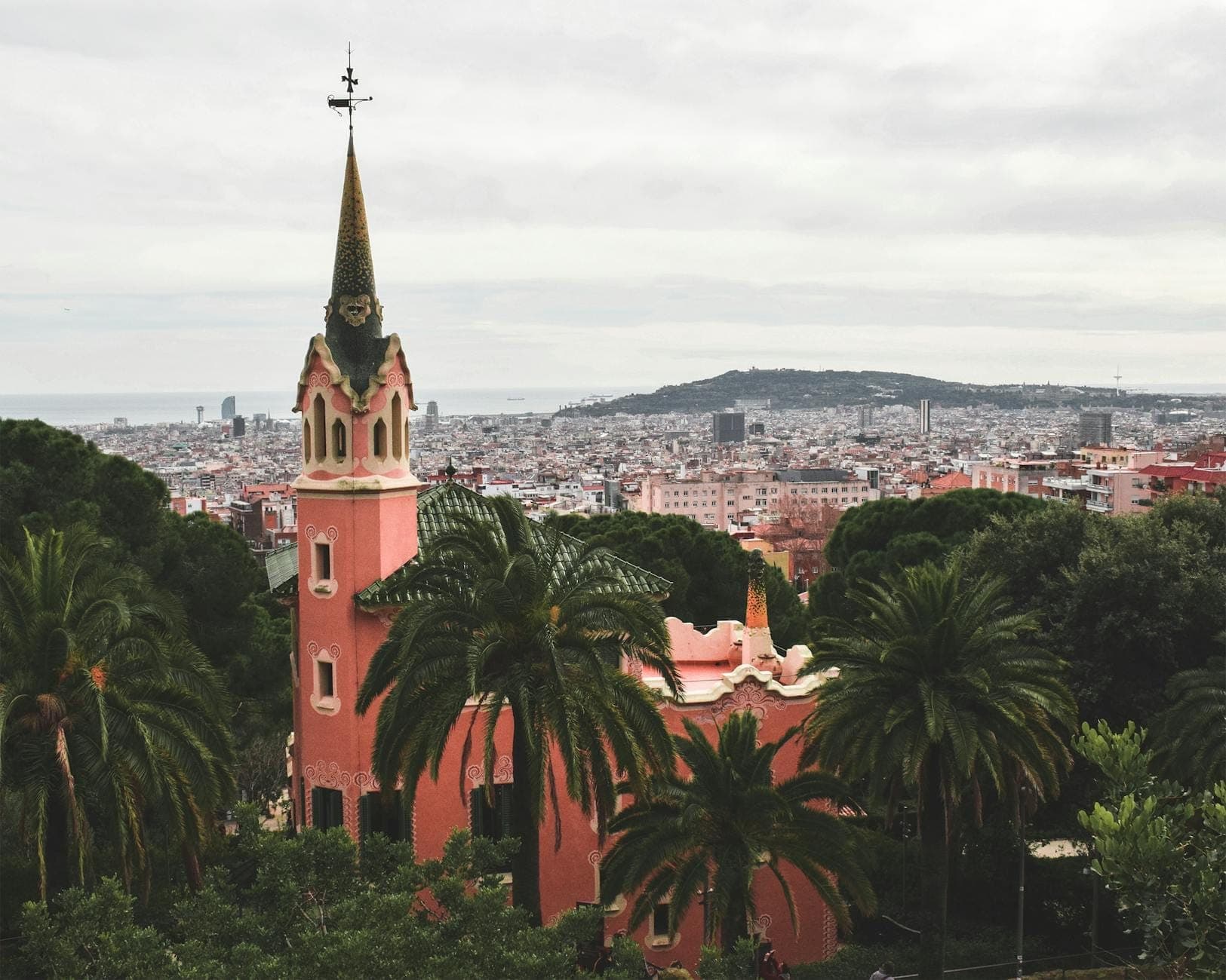 Casa-Museu Gaudí, uma casa neogótica cor-de-rosa com uma torre alta e pontiaguda, cercada por vegetação exuberante e com vista para a cidade de Barcelona.