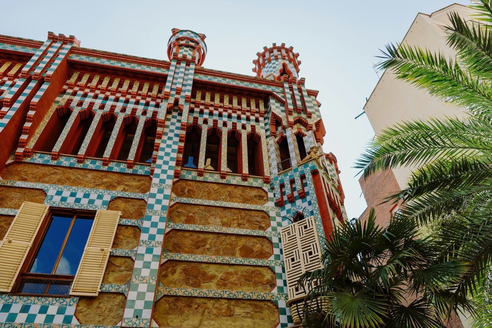 Fachada da Casa Vicens com azulejos de cerâmica coloridos, alvenaria intrincada, janelas arqueadas e palmeiras sob um céu azul limpo.