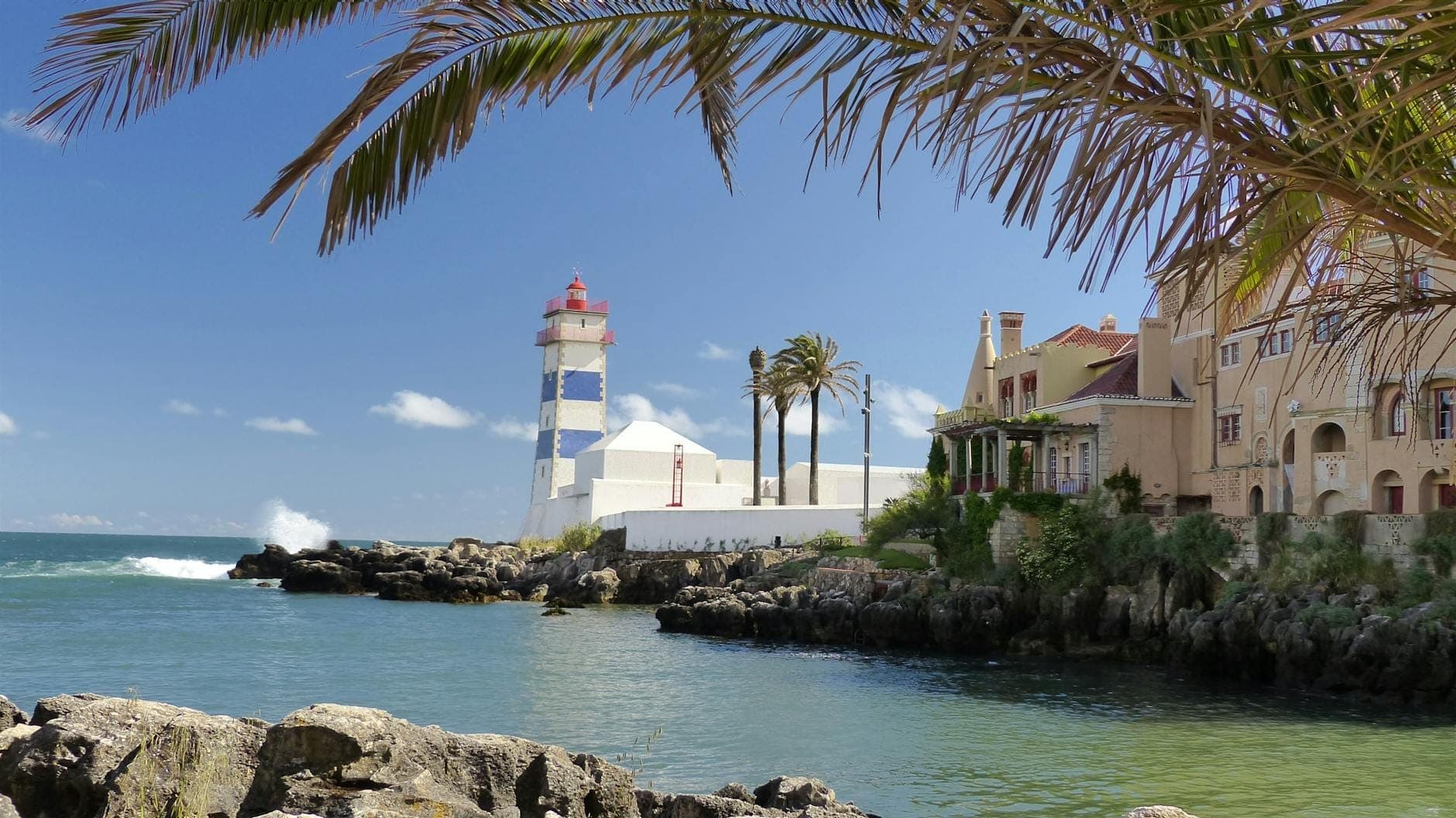 Vista da costa de Cascais com o icônico farol de Santa Marta, elegantes vilas à beira-mar, palmeiras e as águas turquesa do Atlântico em um dia ensolarado.