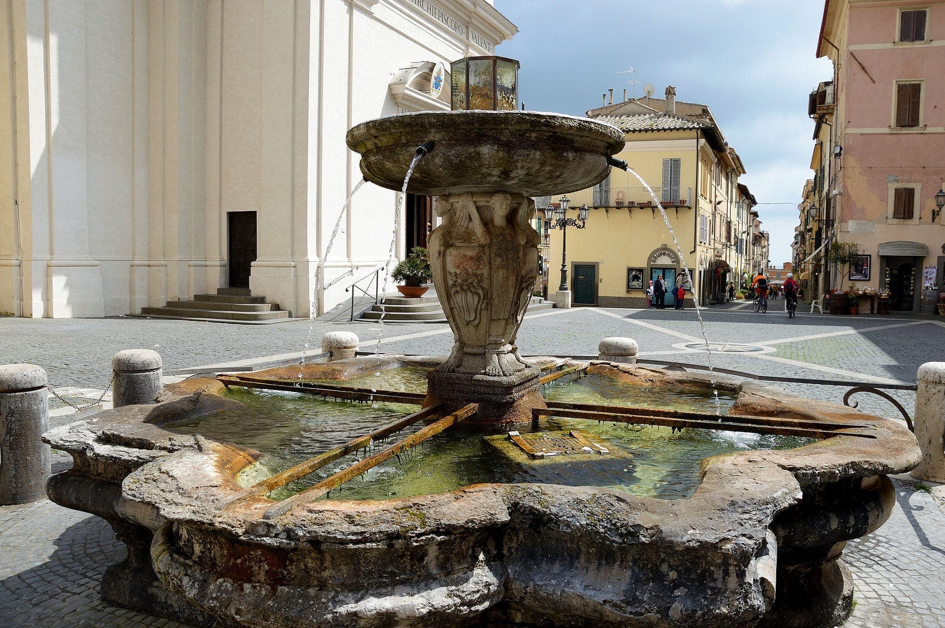 Storica fontana in pietra nella piazza principale di Castel Gandolfo, con acqua che scorre e classici edifici italiani sullo sfondo.