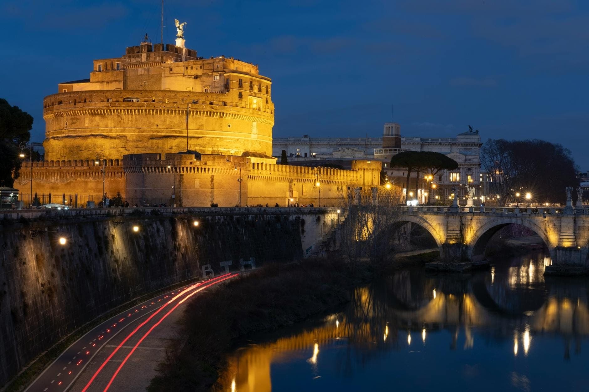 Castel Sant'Angelo lung linh trong ánh đèn đêm, phản chiếu xuống sông Tiber cùng với cây cầu và ánh đèn thành phố tạo nên khung cảnh hoàng hôn huyền ảo.