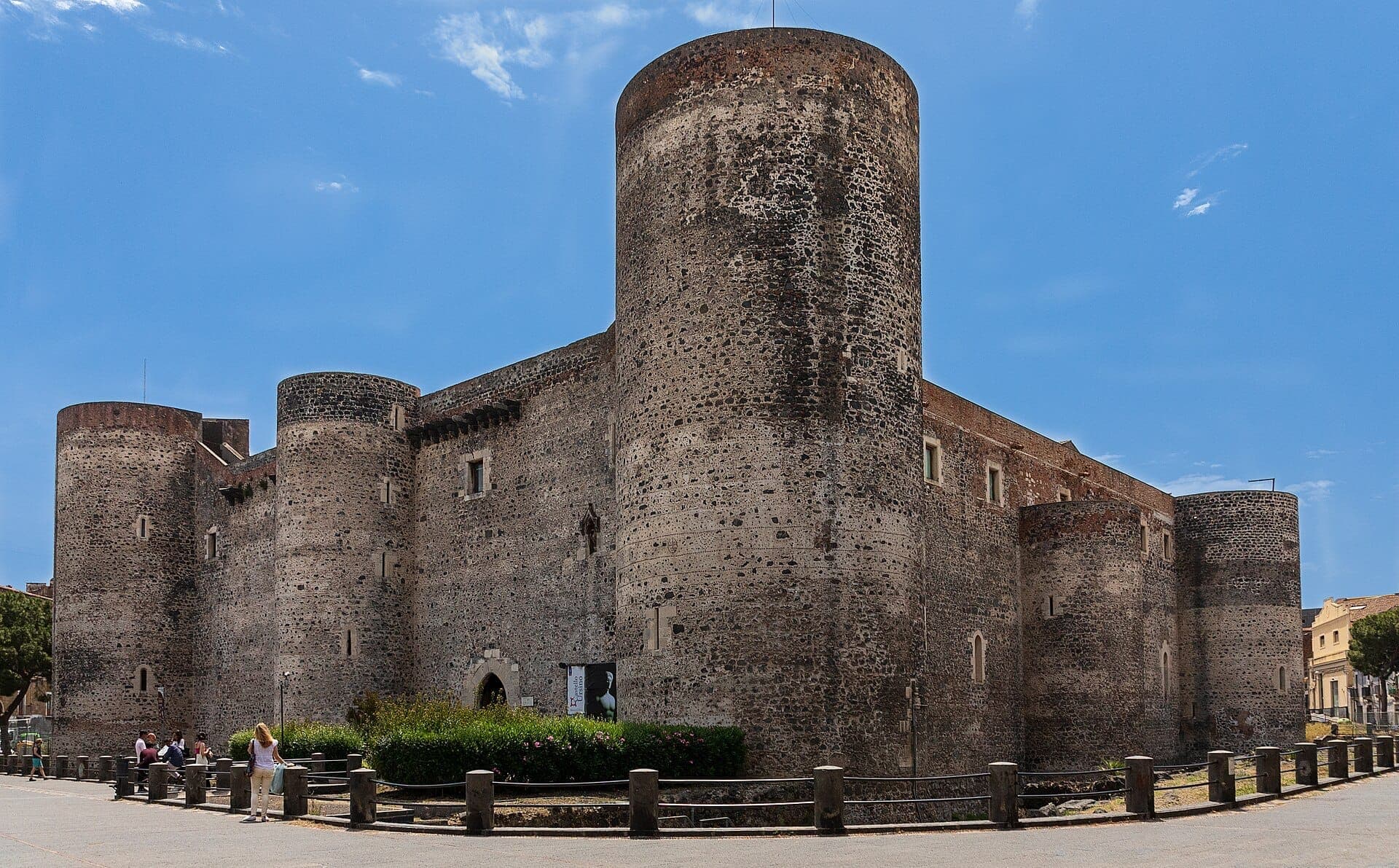 Dinding batu dan menara bulat Castello Ursino di bawah langit biru cerah, dengan pengunjung berjalan dan duduk di dekatnya di Catania.
