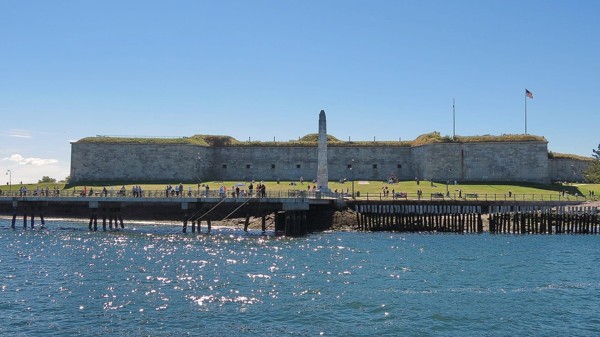 Fort Independence se dresse sur Castle Island avec ses murs en pierre ensoleillés, sa pelouse verdoyante, un monument en obélisque et des visiteurs profitant du front de mer sous un ciel bleu.