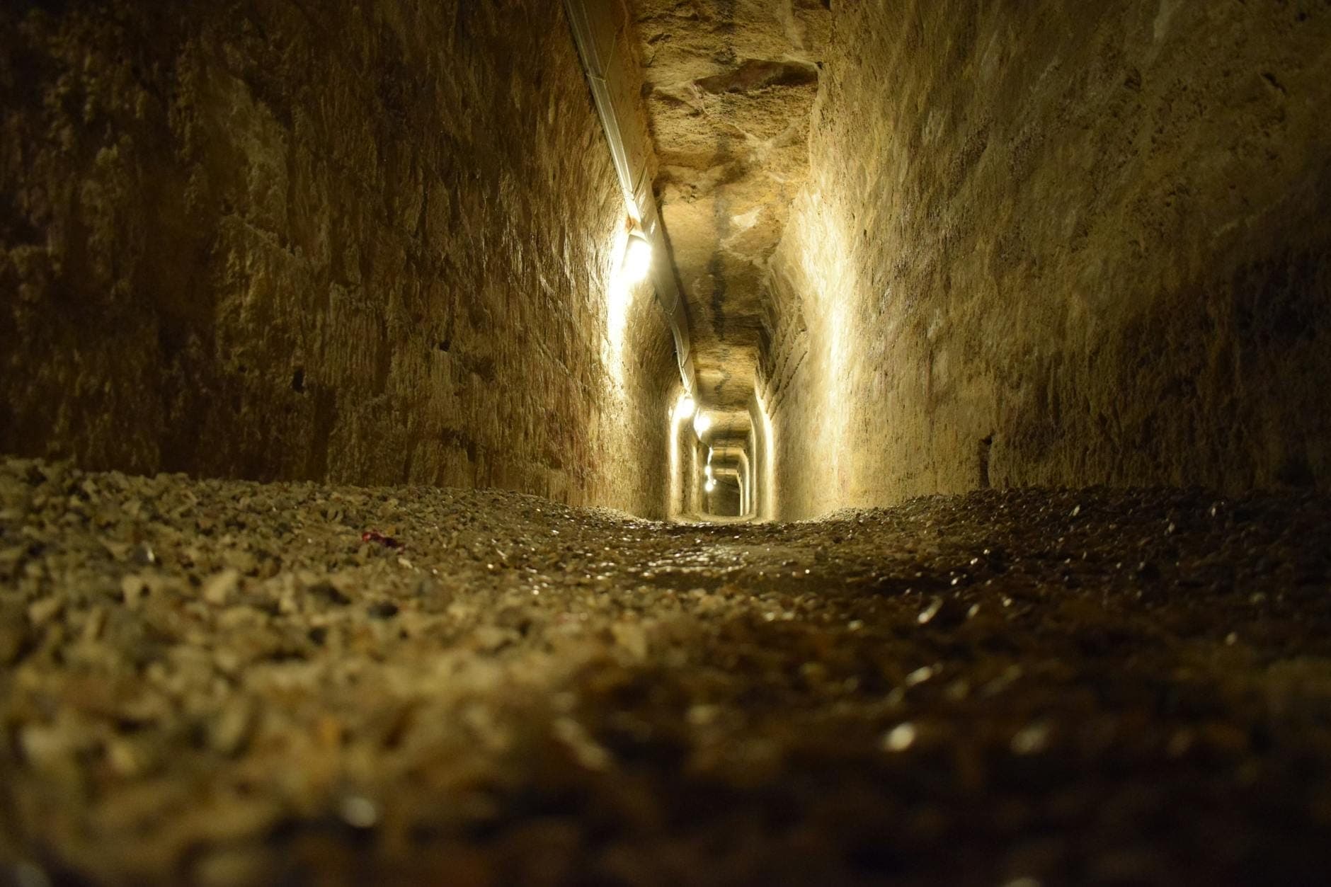 Vista lungo un tunnel sotterraneo in pietra antica, fiocamente illuminato, con pavimento in ghiaia grezza e pareti irregolari: le storiche gallerie delle Catacombe di San Callisto a Roma.