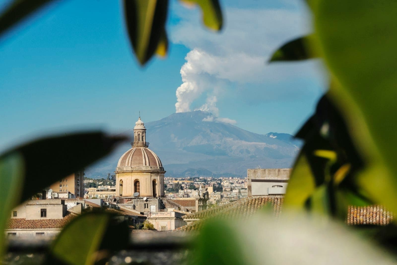 Pemandangan kota Catania dengan kubah barok di latar depan dan Gunung Etna yang mengepulkan asap di latar belakang, dibingkai dedaunan hijau.